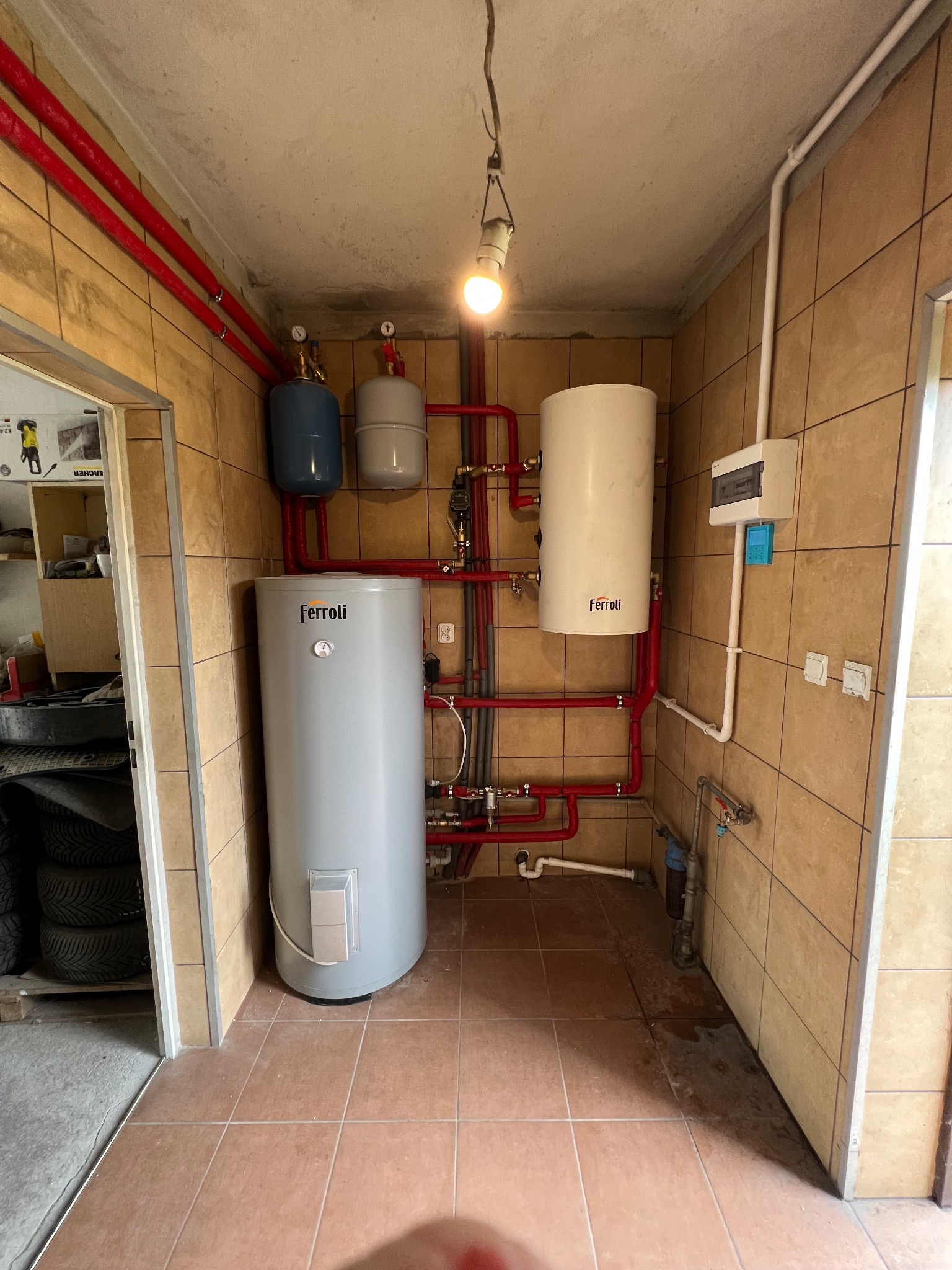 Kotłownia z dwoma zbiornikami Ferroli, instalacją z czerwonych rur, zabezpieczeniami i rozdzielnicą elektryczną na beżowych płytkach, oświetlona wiszącą żarówką.