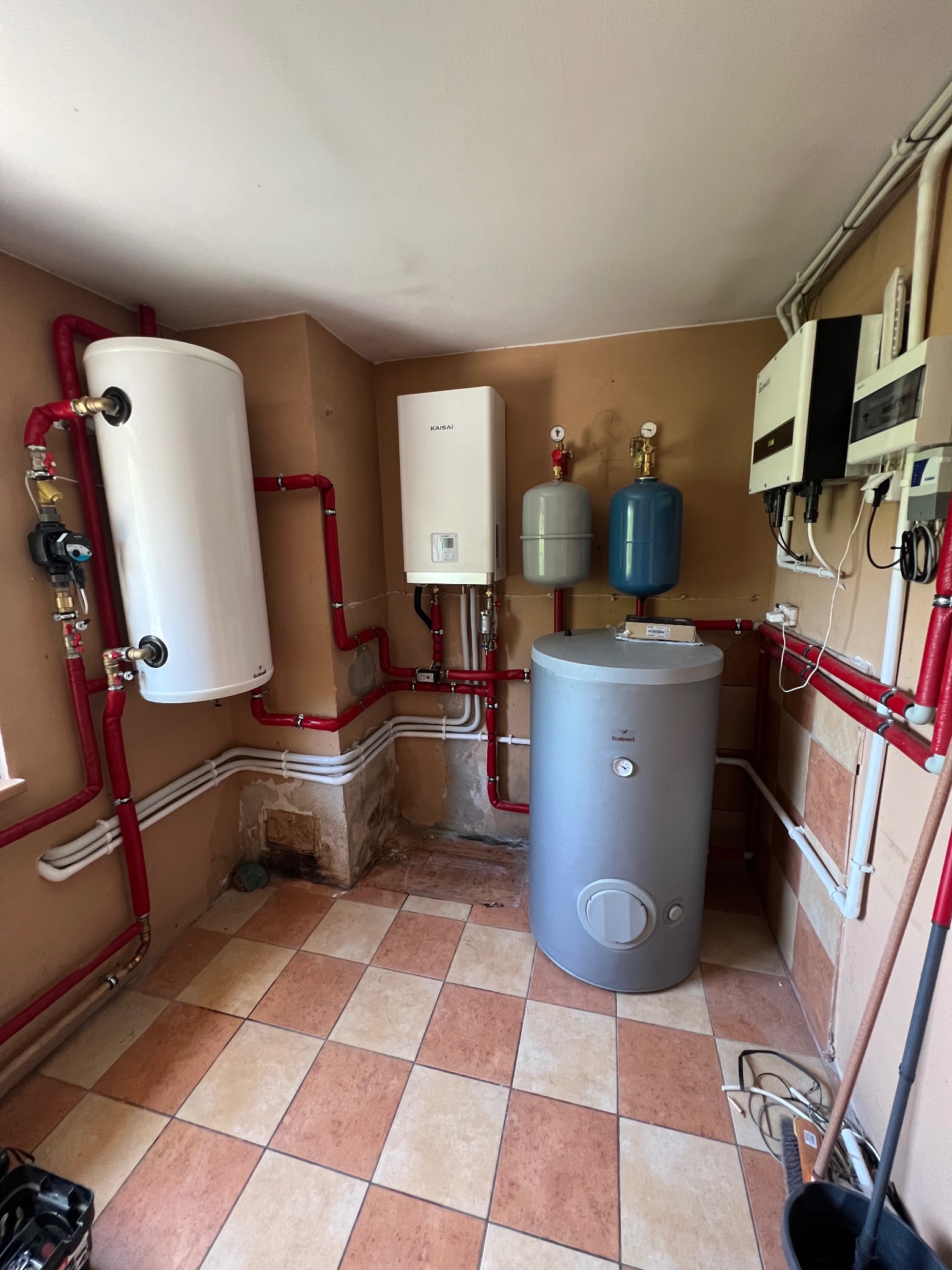 Kotłownia z zainstalowanym białym zasobnikiem ciepłej wody, piecem gazowym Kaisai, szarym bojlerem Galmet i dwoma małymi zbiornikami wyrównawczymi, połączonymi rurami w kolorze czerwonym.