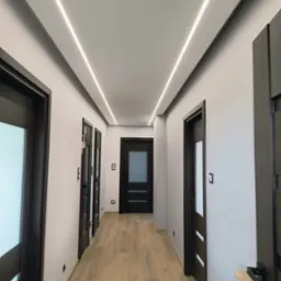 Nowoczesny korytarz z oświetleniem LED w suficie, ciemne drzwi z przeszkleniami, jasne panele podłogowe i szare ściany.