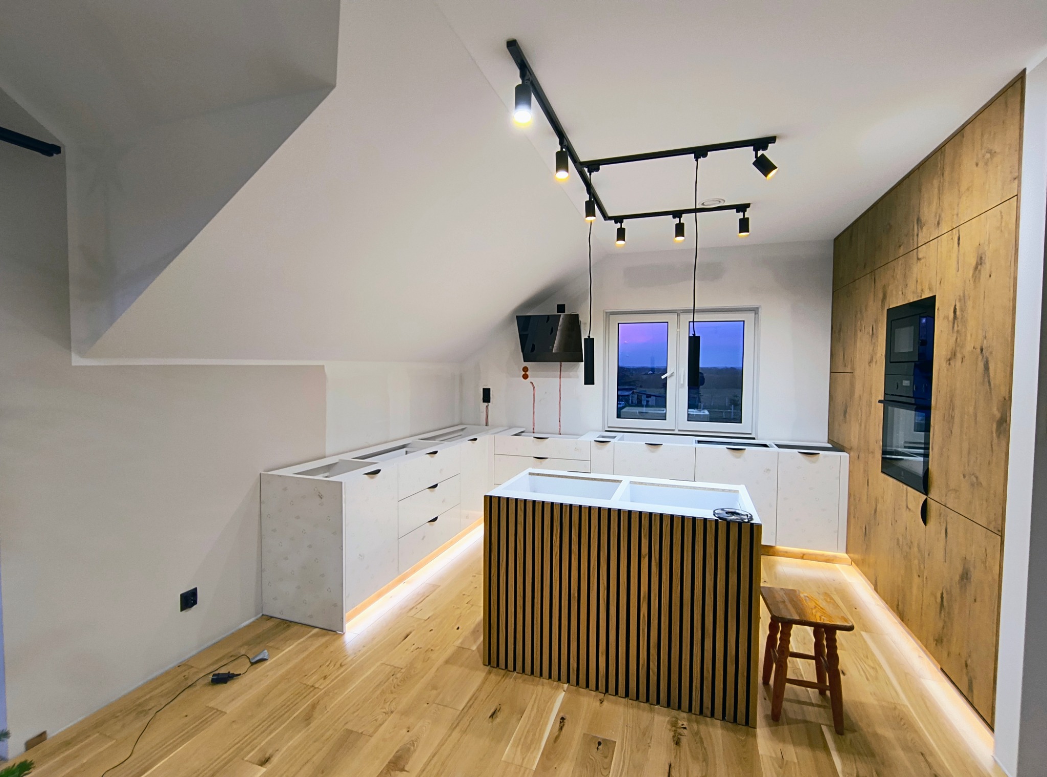 Nowoczesna kuchnia w trakcie montażu: białe szafki z podświetleniem LED, drewniany panel ścienny z piekarnikiem, wyspa z pionowych listew, oświetlenie szynowe na suficie pod skosem.