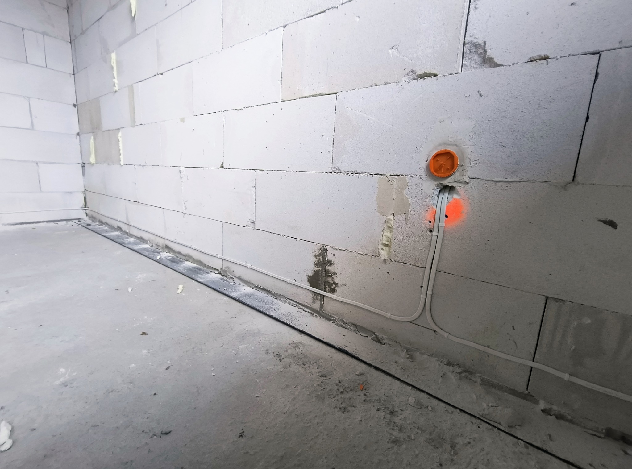 Wnętrze w Szczukowicach w trakcie prac elektrycznych: widoczna puszka instalacyjna z pomarańczowym deklem, kable elektryczne prowadzone wzdłuż ściany z betonu komórkowego, oznaczone pomarańczowym...