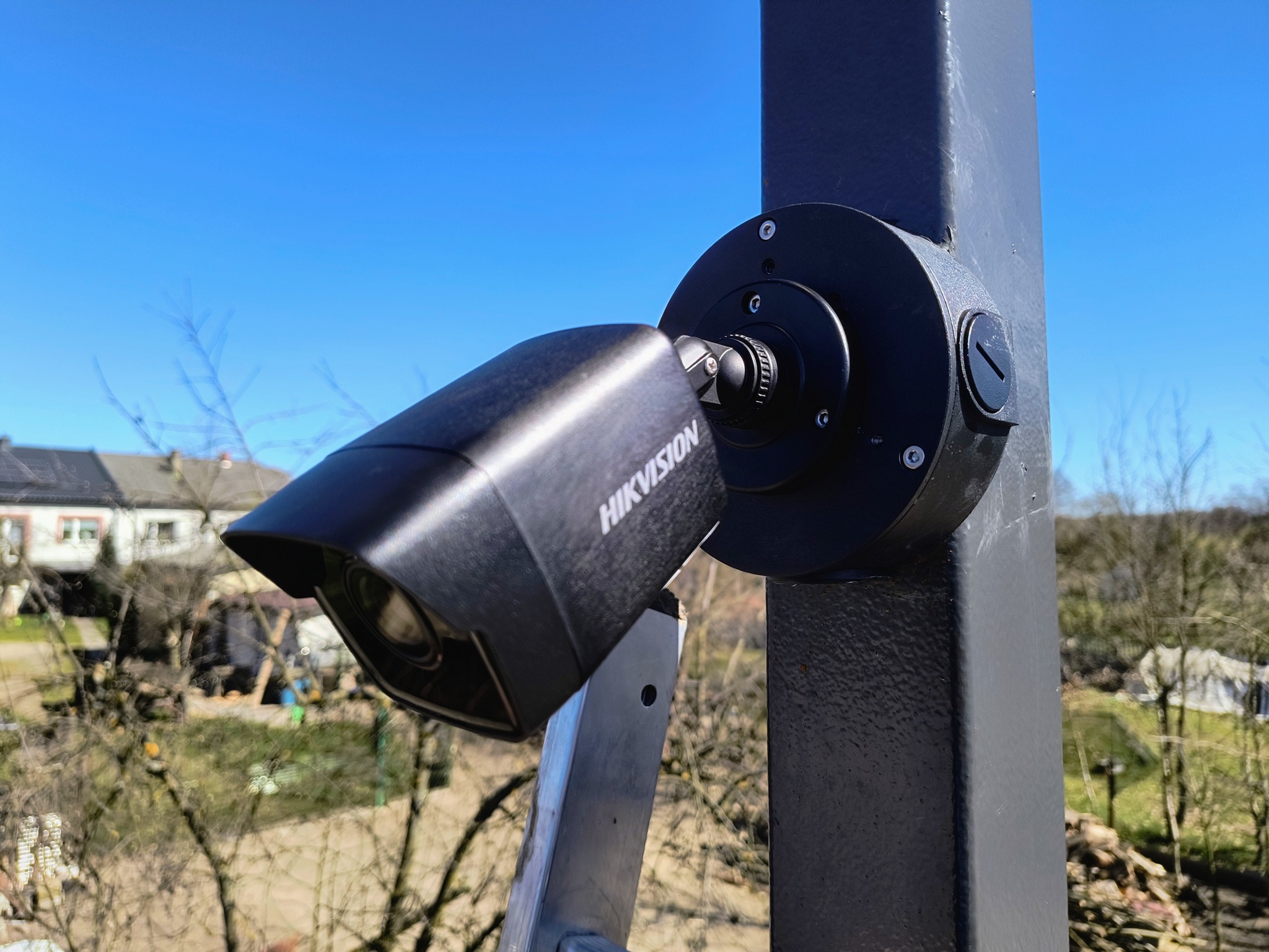 Czarna kamera monitoringu Hikvision zamontowana na metalowym słupku, widok z dołu, na tle błękitnego nieba i zabudowań.