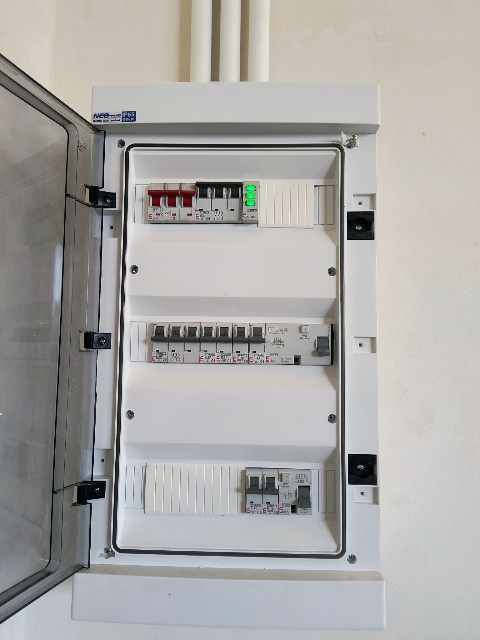 Otwarta skrzynka elektryczna z widocznymi bezpiecznikami, wyłącznikami różnicowoprądowymi i listwą z zielonymi diodami, zamontowana na białej ścianie.