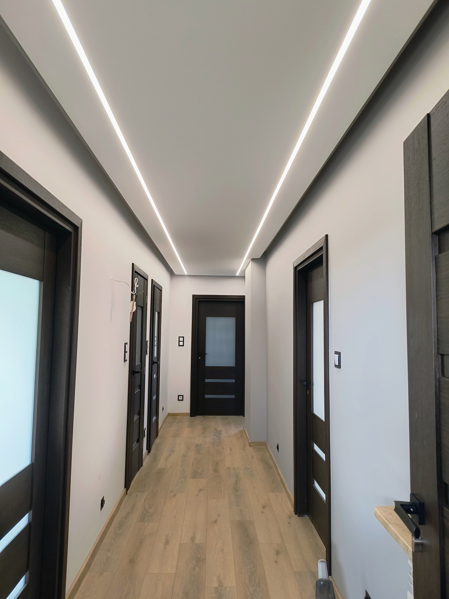 Nowoczesny korytarz z oświetleniem LED w suficie, ciemne drzwi z przeszkleniami, jasne panele podłogowe i szare ściany.