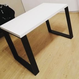 Stal fox - Nowoczesny stół z białym blatem i czarnymi, metalowymi nogami o geometrycznym kształcie, ustawiony na podłodze z paneli imitujących drewno.