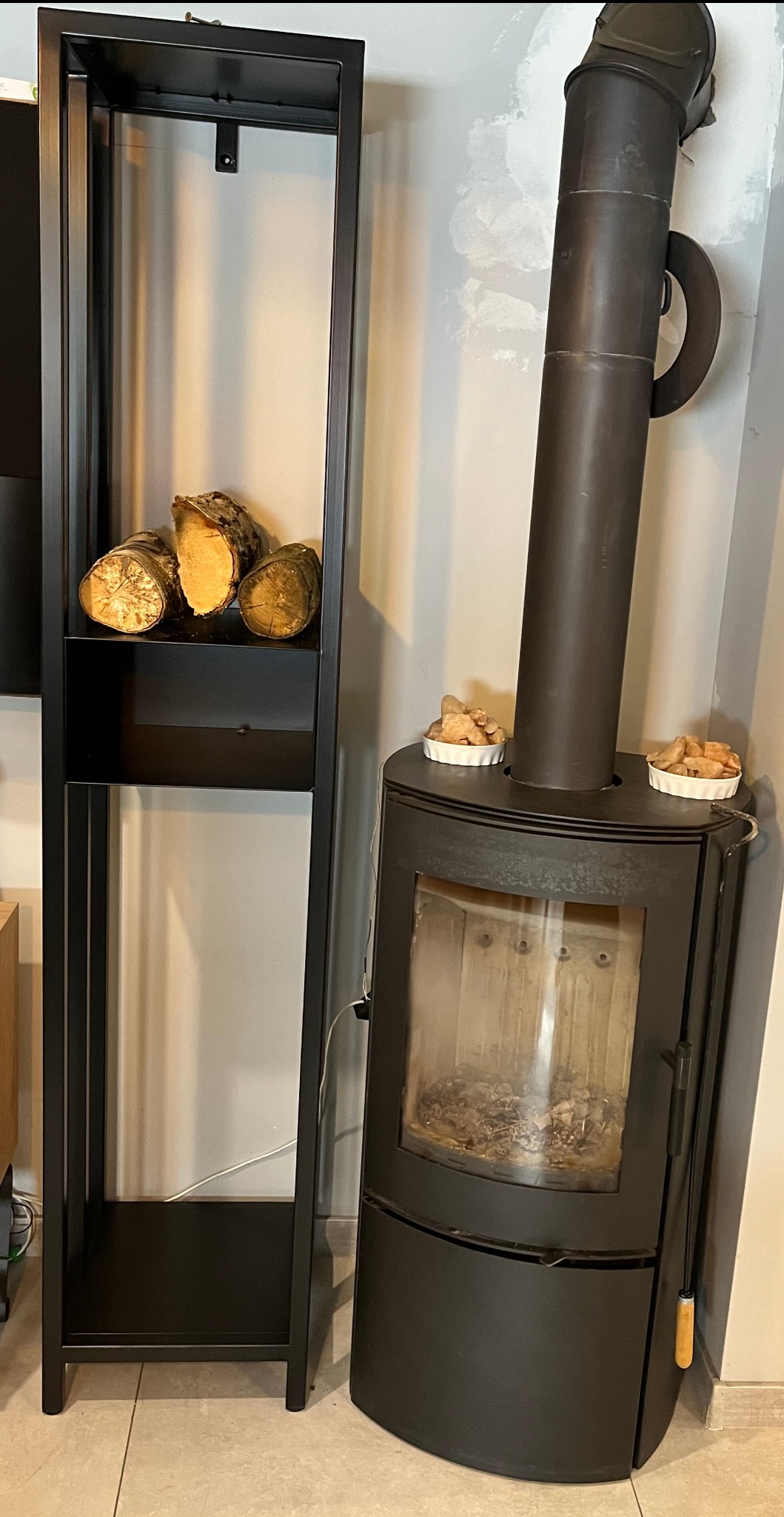 Czarny piec wolnostojący typu koza z rurą odprowadzającą dym, obok czarny regał z ułożonymi kawałkami drewna na półce, na piecu dwie małe miseczki z brykietami do rozpałki.