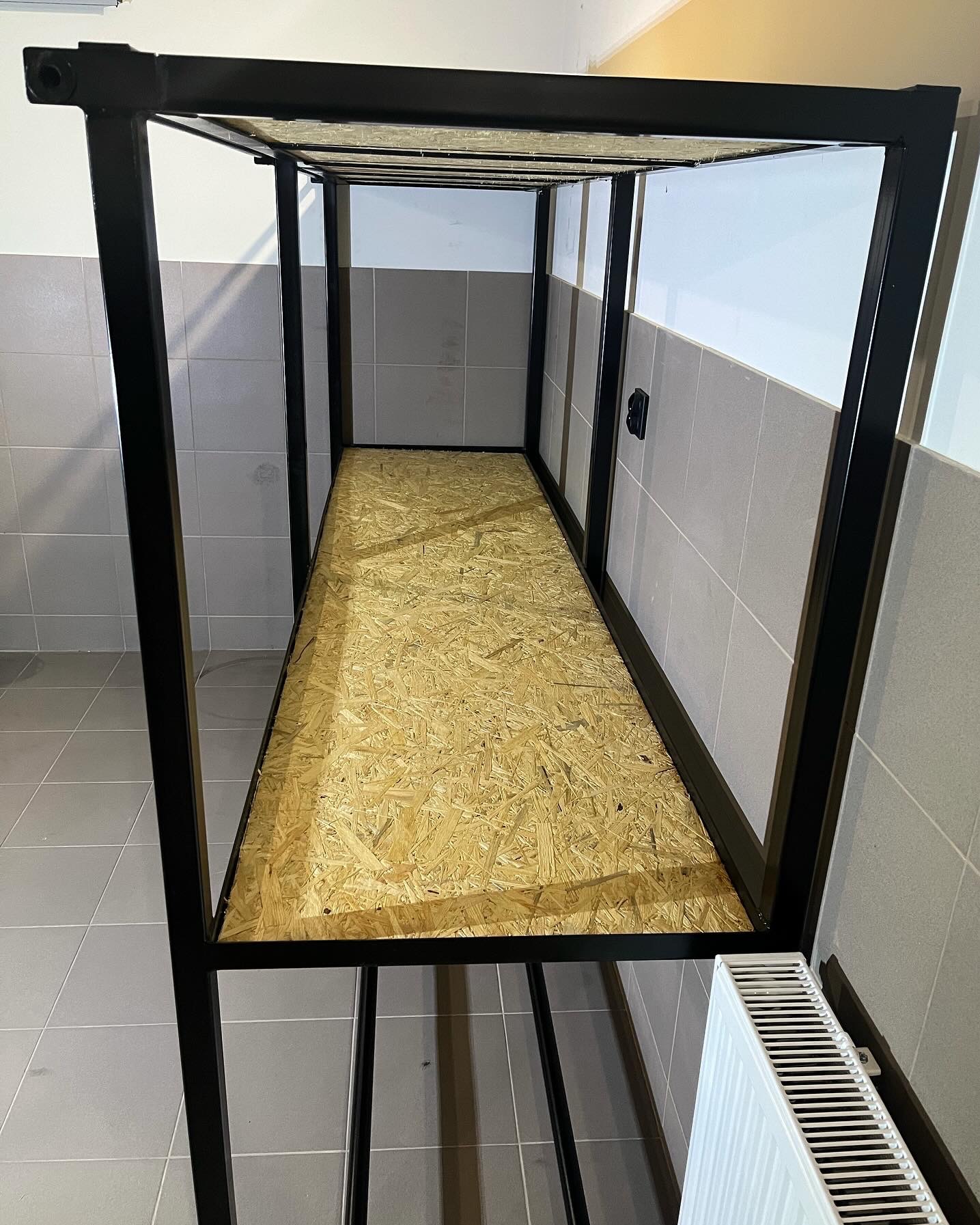 Wnętrze metalowej konstrukcji z wypełnieniem z płyty OSB, widoczne na tle wykafelkowanej ściany i podłogi, z grzejnikiem u dołu kadru.