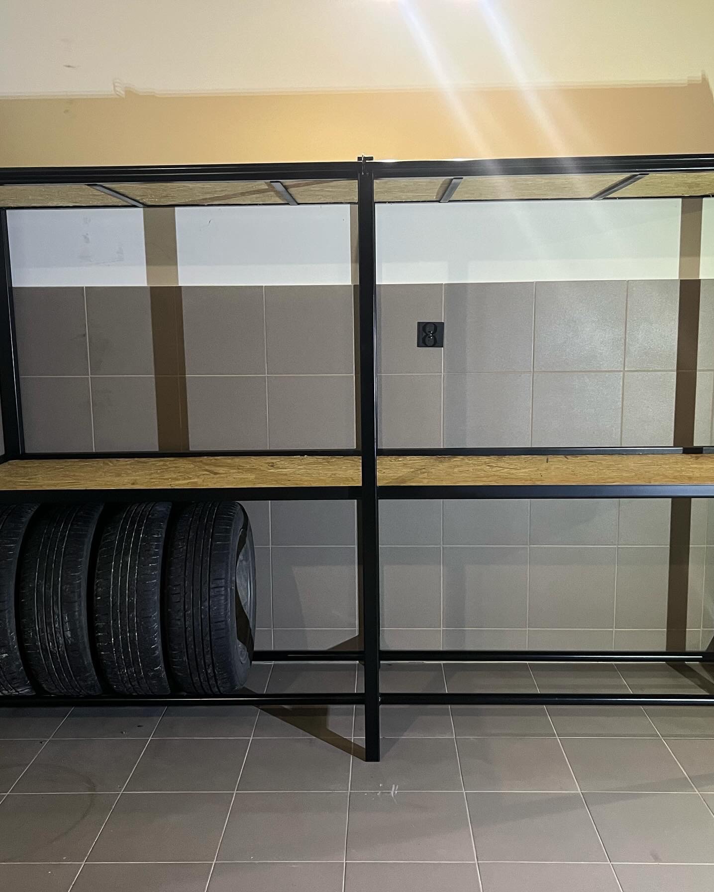 Metalowy regał z drewnianymi półkami, na których leżą opony samochodowe, ustawiony w pomieszczeniu wyłożonym szarymi płytkami.