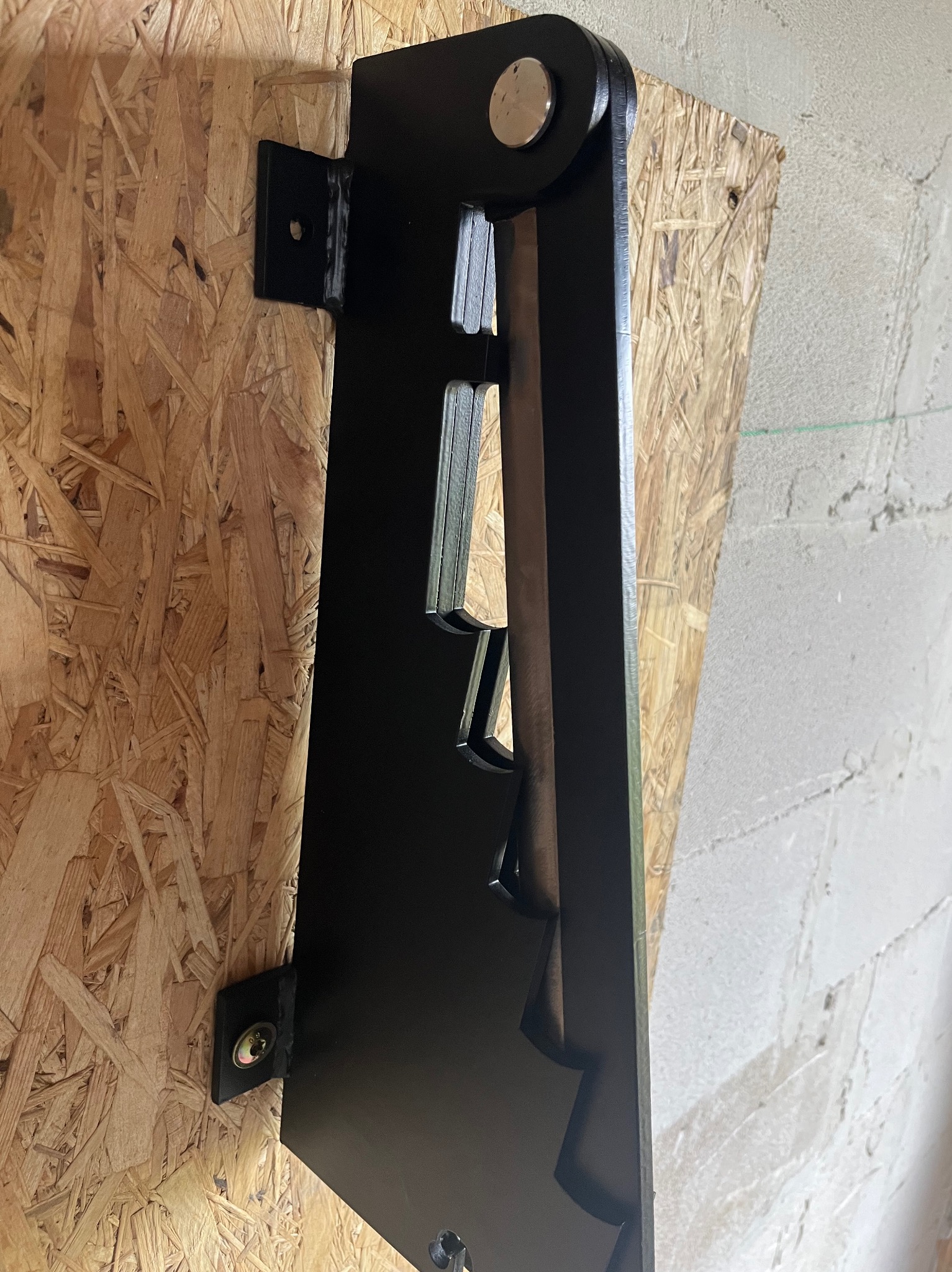 Czarny, metalowy element konstrukcyjny z wyciętymi kształtami, zamocowany do płyty OSB i ściany, widoczny z boku.