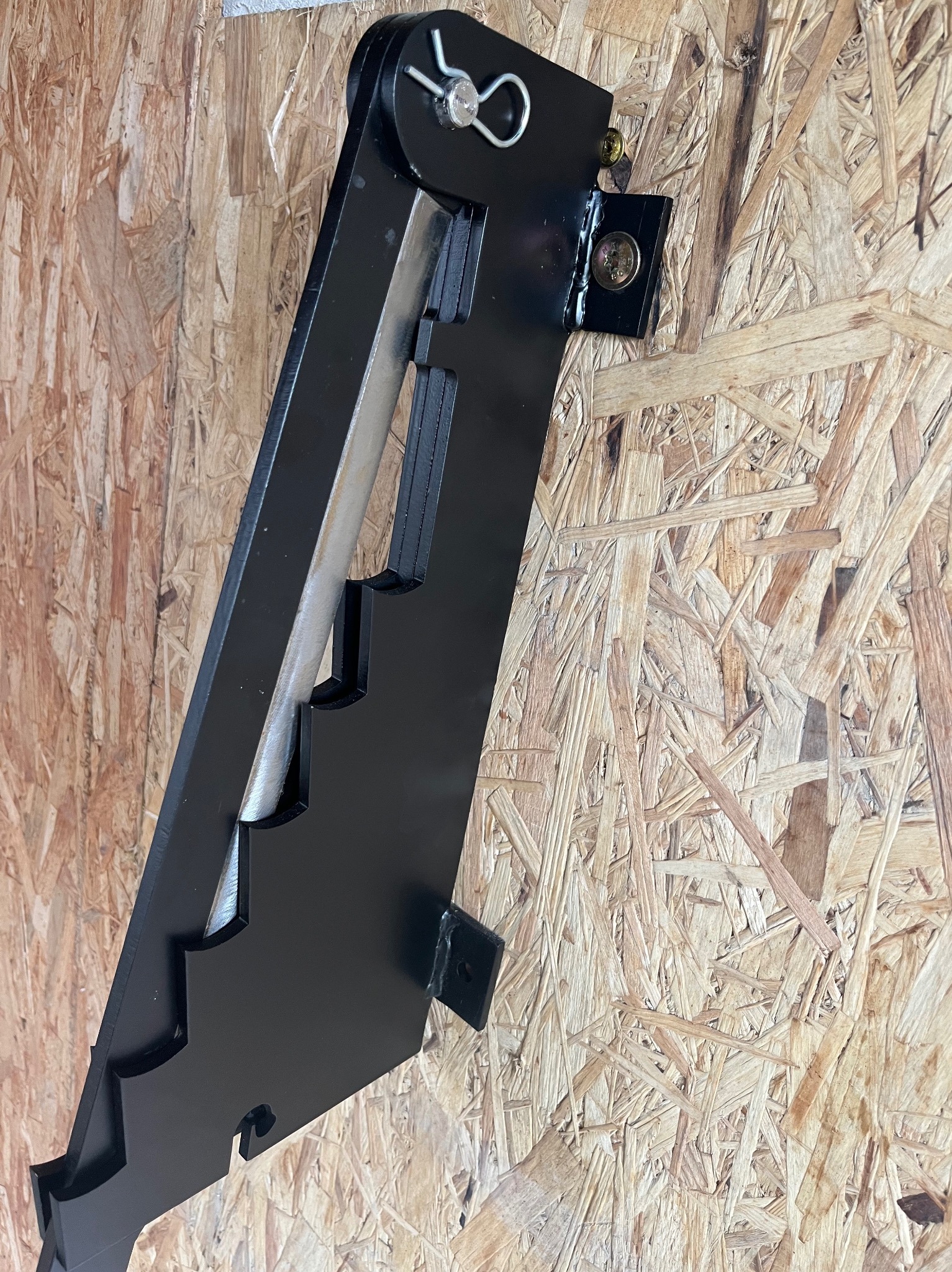 Czarny, metalowy element konstrukcyjny z wycięciami, przymocowany do płyty OSB za pomocą śrub i zawleczki, widoczne detale mocowania.