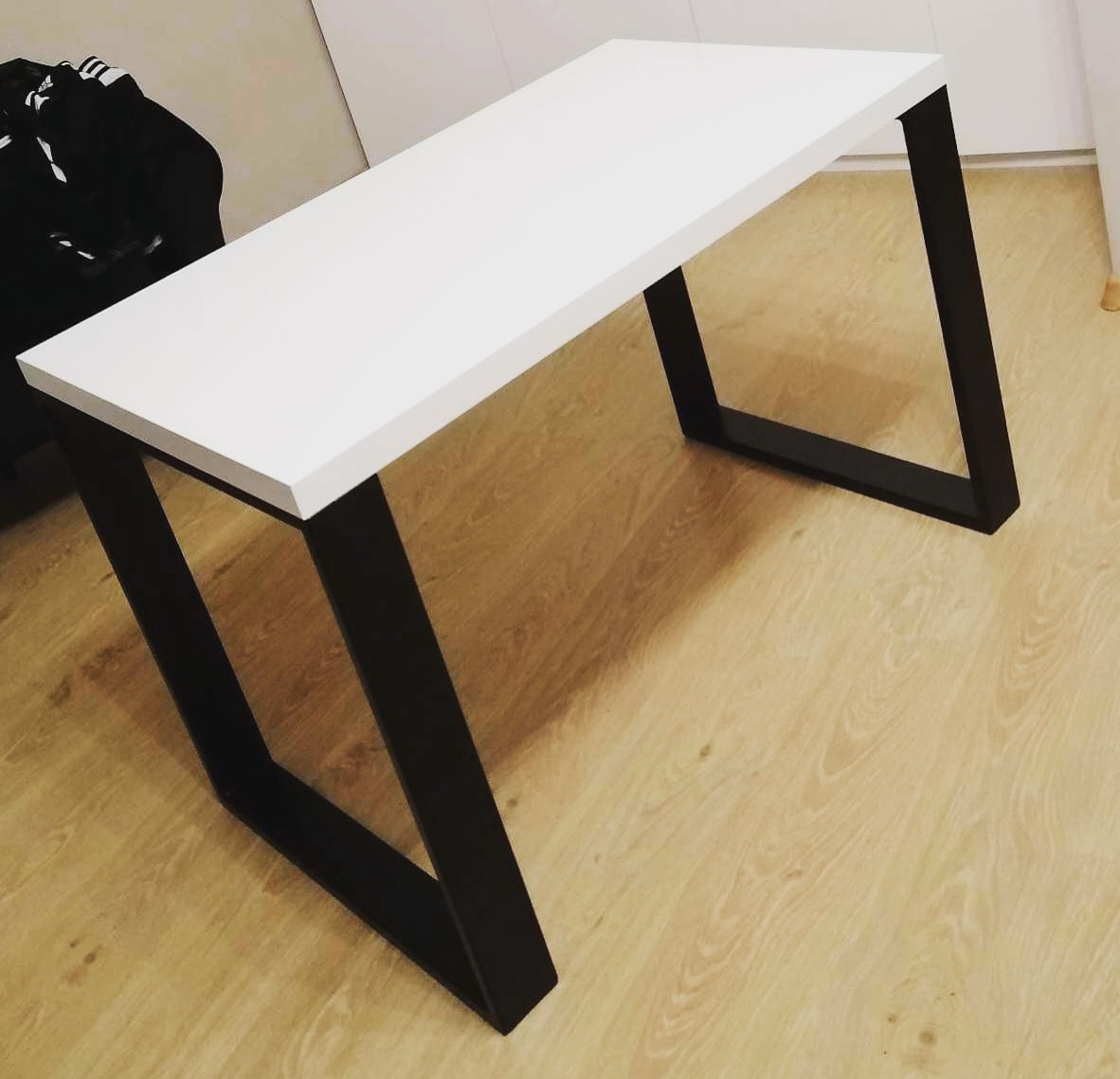 Nowoczesny stół z białym blatem i czarnymi, metalowymi nogami o geometrycznym kształcie, ustawiony na podłodze z paneli imitujących drewno.