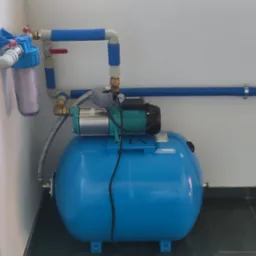 Hydrofor z filtrem wody i rurami w niebieskiej izolacji, zamontowany w pomieszczeniu z szarymi płytkami podłogowymi i białą ścianą.