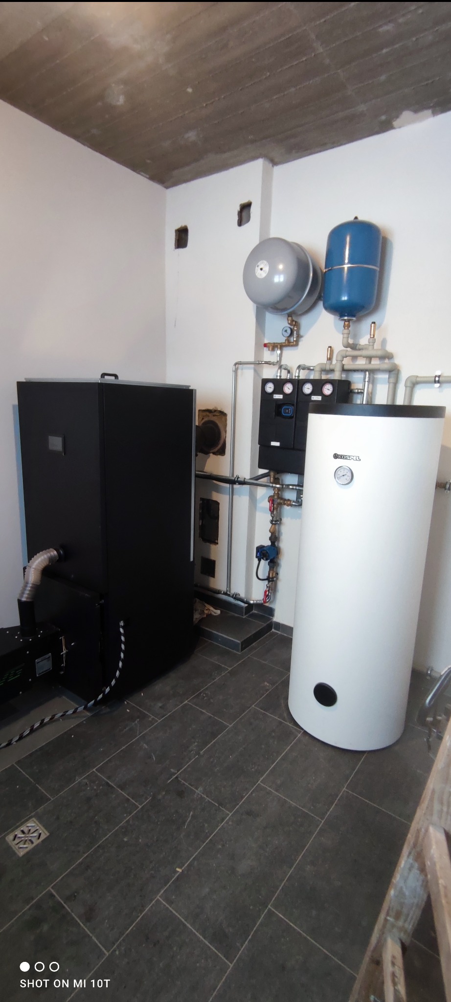 Kotłownia z kotłem na paliwo stałe, zbiornikiem buforowym i osprzętem, widoczne rury i zawory, surowy betonowy sufit, ciemna podłoga