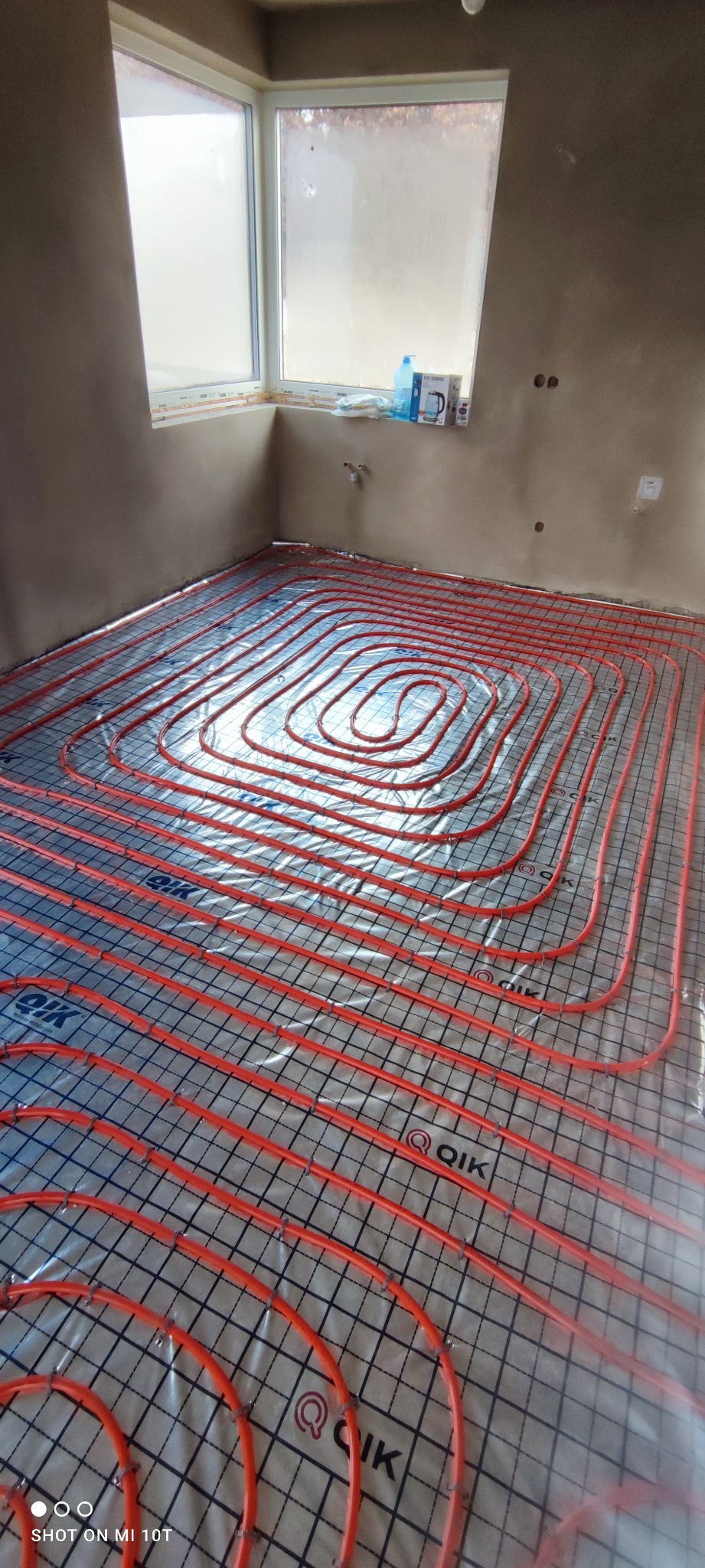 Czerwone rury ogrzewania podłogowego ułożone na folii z nadrukiem siatki w surowym pomieszczeniu z oknem, widoczne akcesoria instalacyjne.