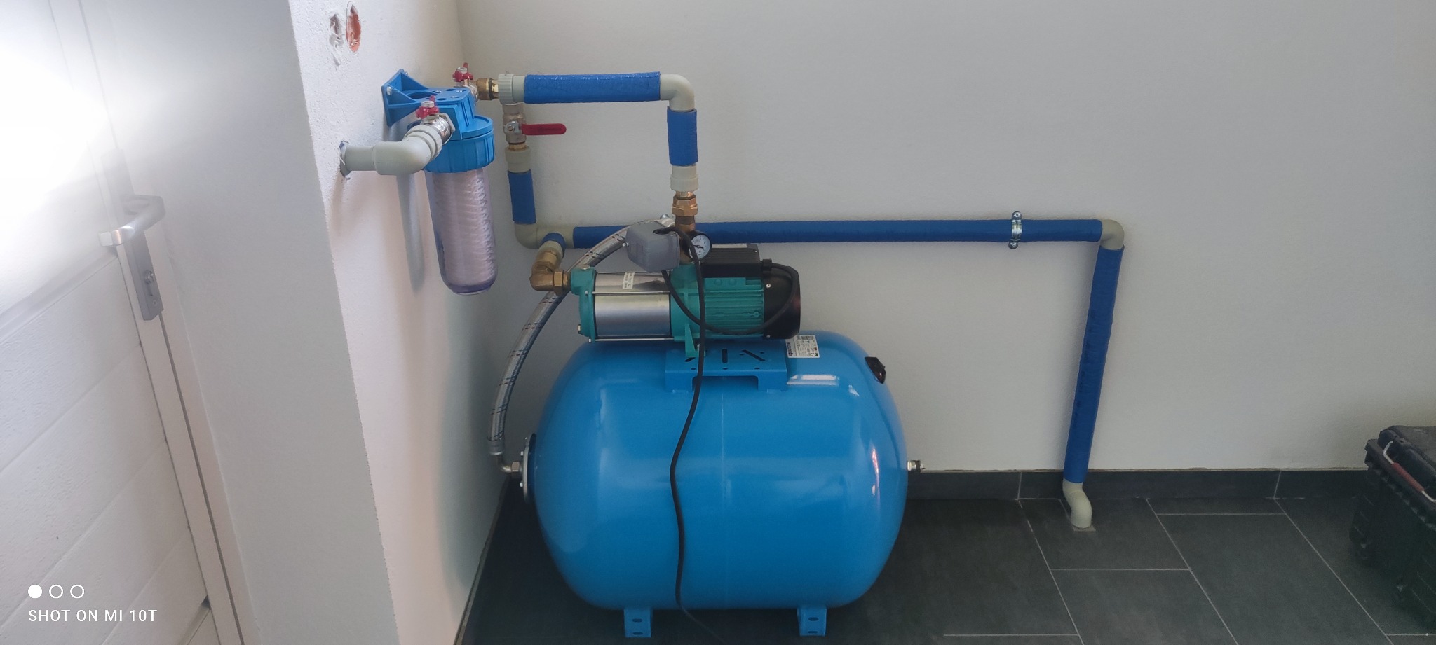 Hydrofor z filtrem wody i rurami w niebieskiej izolacji, zamontowany w pomieszczeniu z szarymi płytkami podłogowymi i białą ścianą.