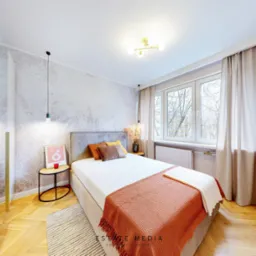 Jasna sypialnia z łóżkiem, drewnianą podłogą w jodełkę i tapetą w delikatne wzory, oświetlona wiszącymi lampami i lampą sufitową, z widokiem na drzewa za oknem.