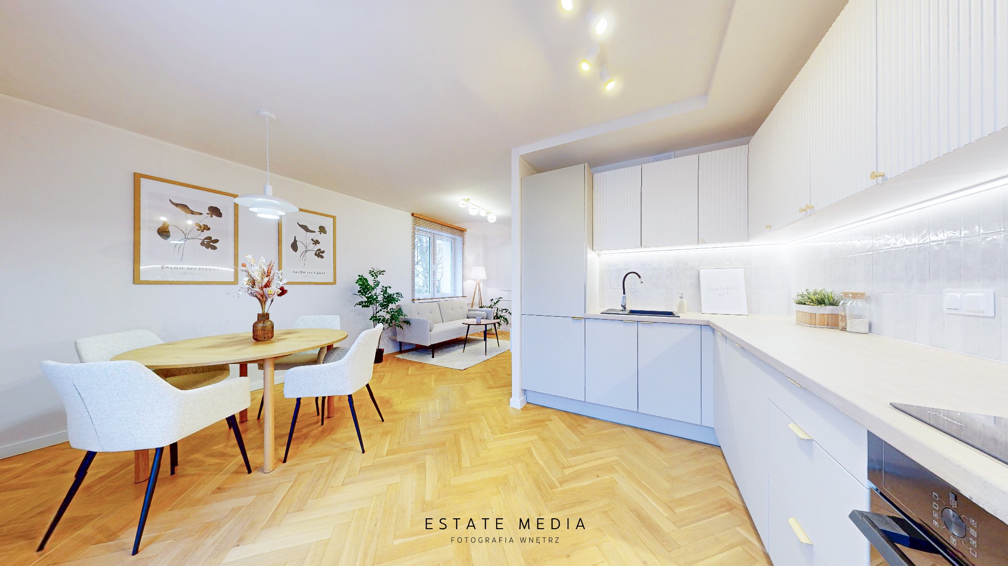 Jasne, przestronne wnętrze mieszkania z parkietem w jodełkę, łączące kuchnię z jadalnią i salonem. Widoczne białe meble kuchenne z subtelnym oświetleniem LED, okrągły stół z krzesłami...