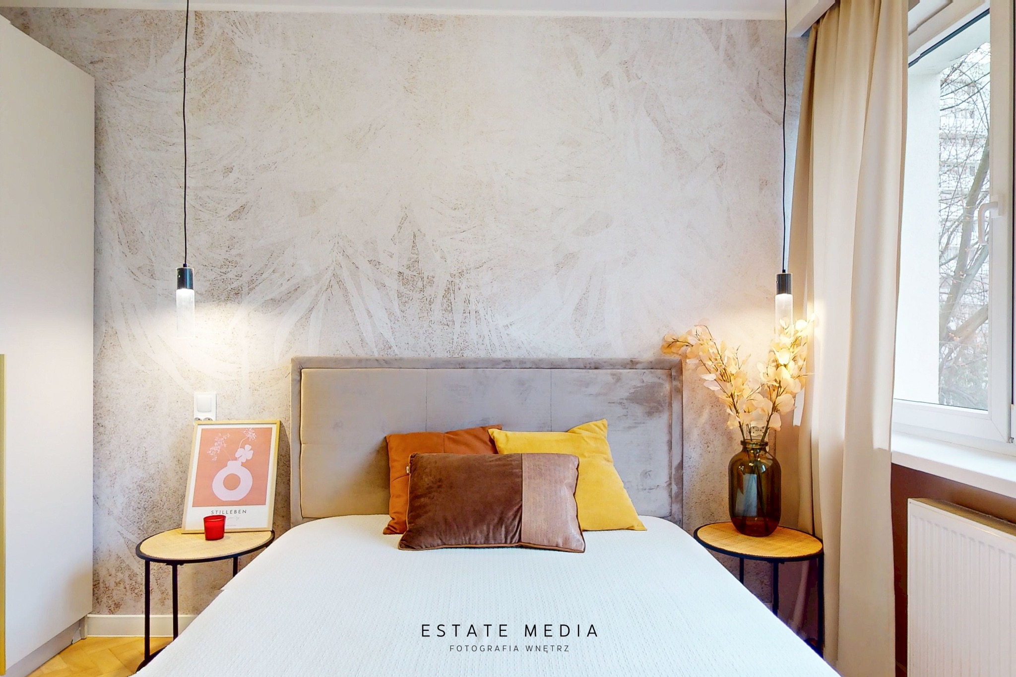 Jasna sypialnia z wzorzystą tapetą, beżowym zagłówkiem, kolorowymi poduszkami i wiszącymi lampami po bokach łóżka. Na stoliku nocnym plakat i czerwona świeczka.