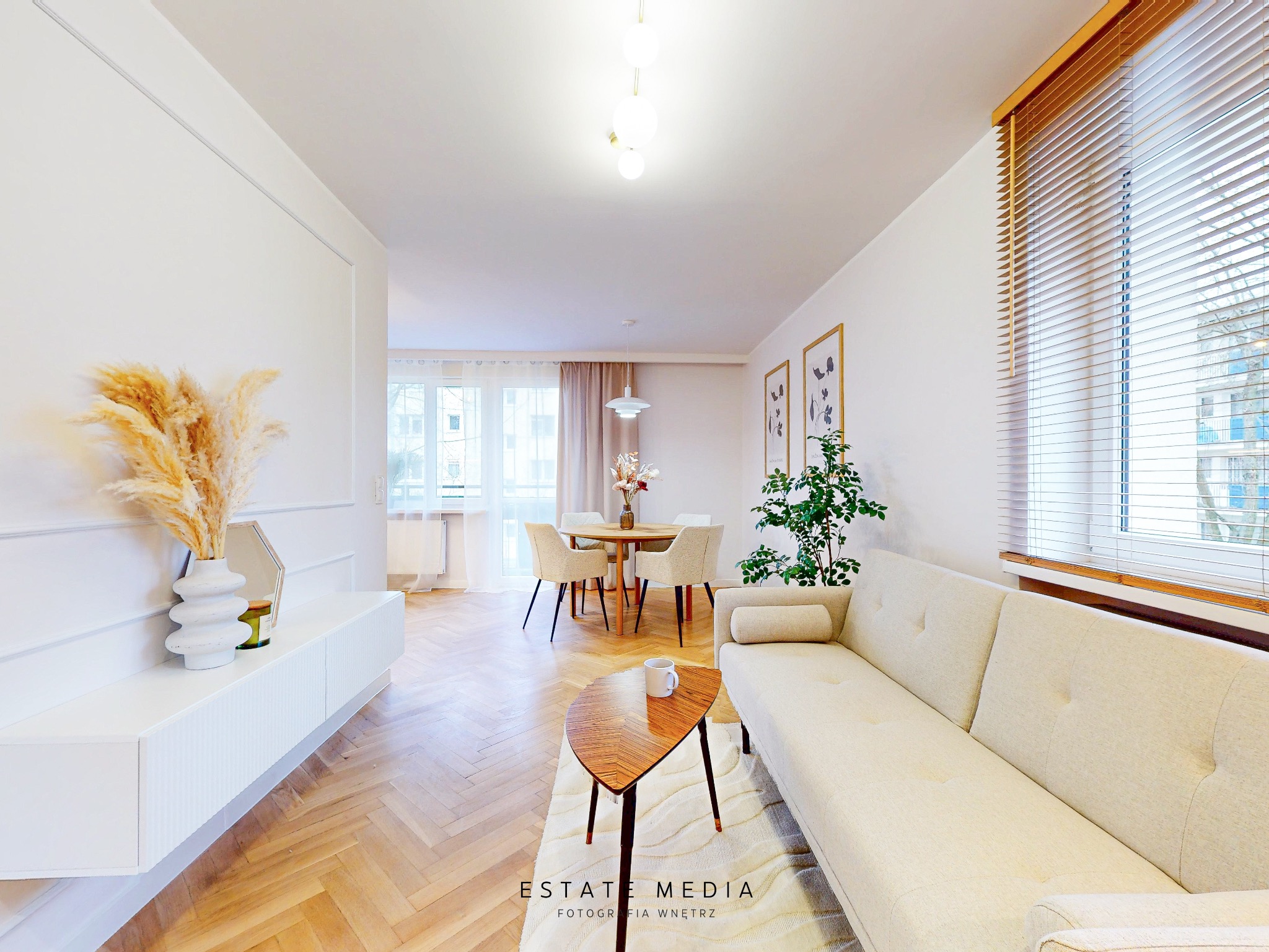 Jasne, przestronne wnętrze mieszkania z parkietem w jodełkę, beżową sofą, drewnianym stolikiem kawowym, okrągłym stołem jadalnym i dekoracjami z suszonych traw w wazonie.