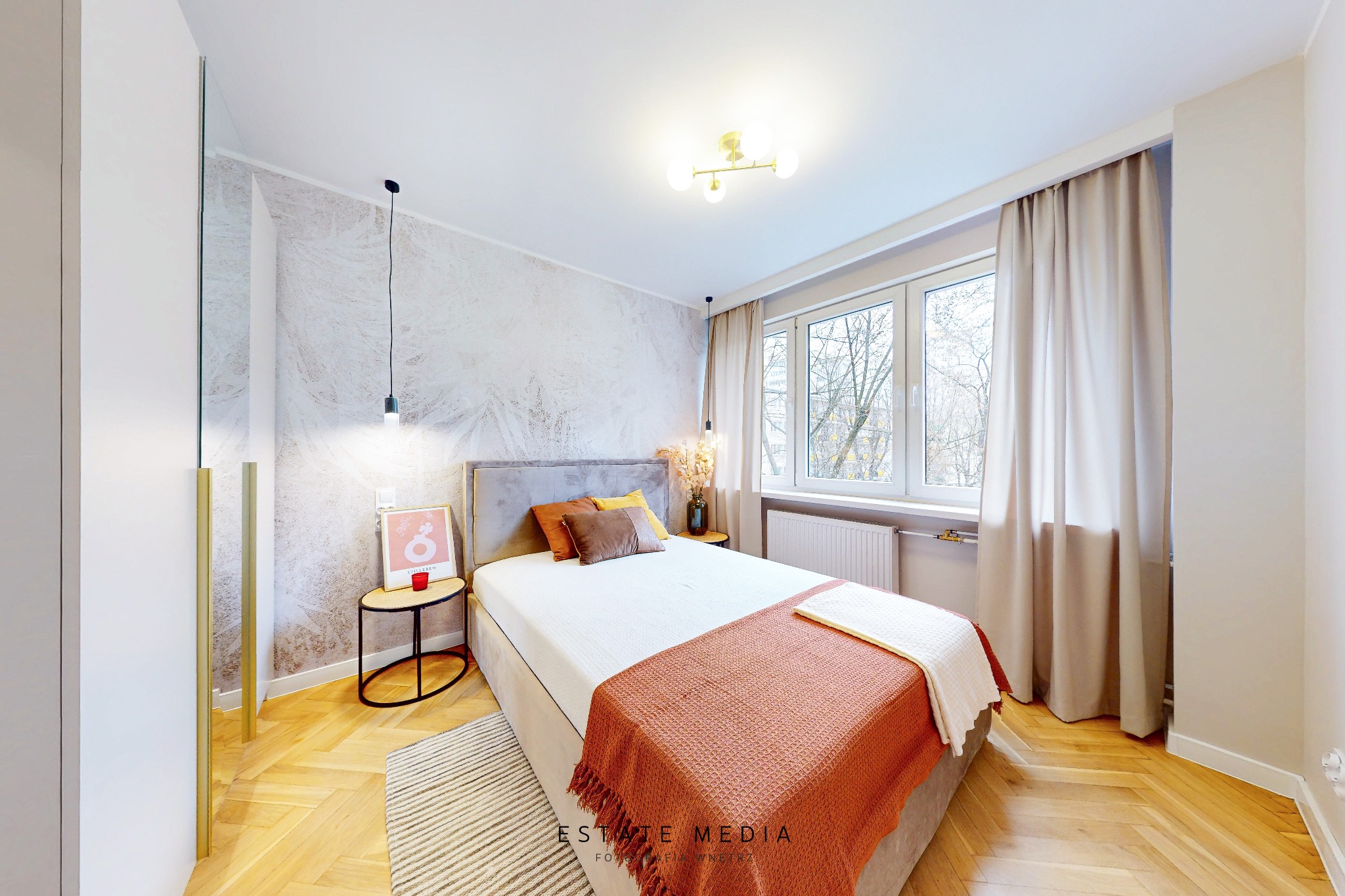 Jasna sypialnia z łóżkiem, drewnianą podłogą w jodełkę i tapetą w delikatne wzory, oświetlona wiszącymi lampami i lampą sufitową, z widokiem na drzewa za oknem.