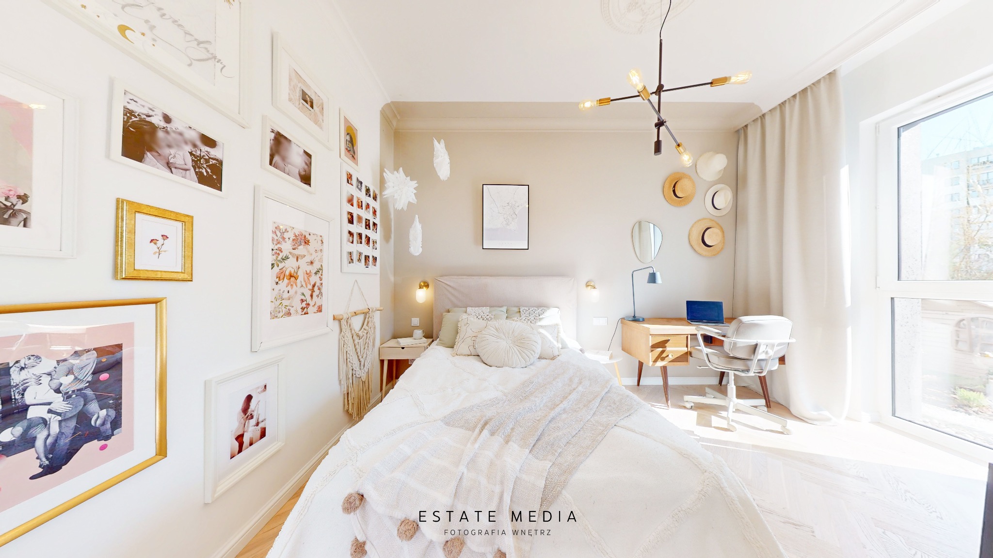 Jasna sypialnia z łóżkiem, biurkiem i galerią obrazów na ścianie, oświetlona naturalnym światłem wpadającym przez duże okno.