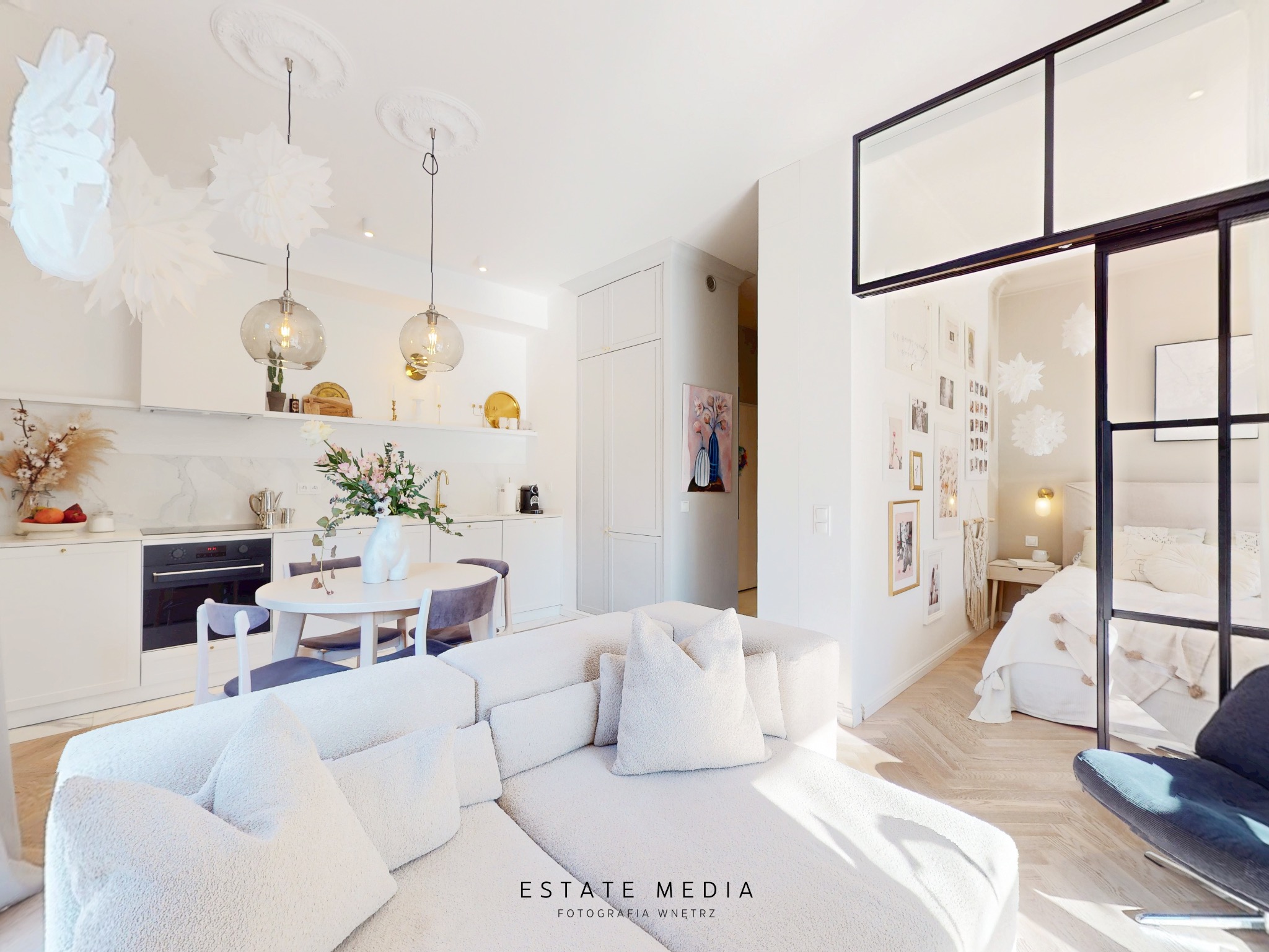 Jasne, przestronne wnętrze mieszkania z otwartą kuchnią, jadalnią i sypialnią oddzieloną szklaną ścianką, z przewagą bieli i naturalnego światła, z dekoracyjnymi lampami, kwiatami i tekstyliami.