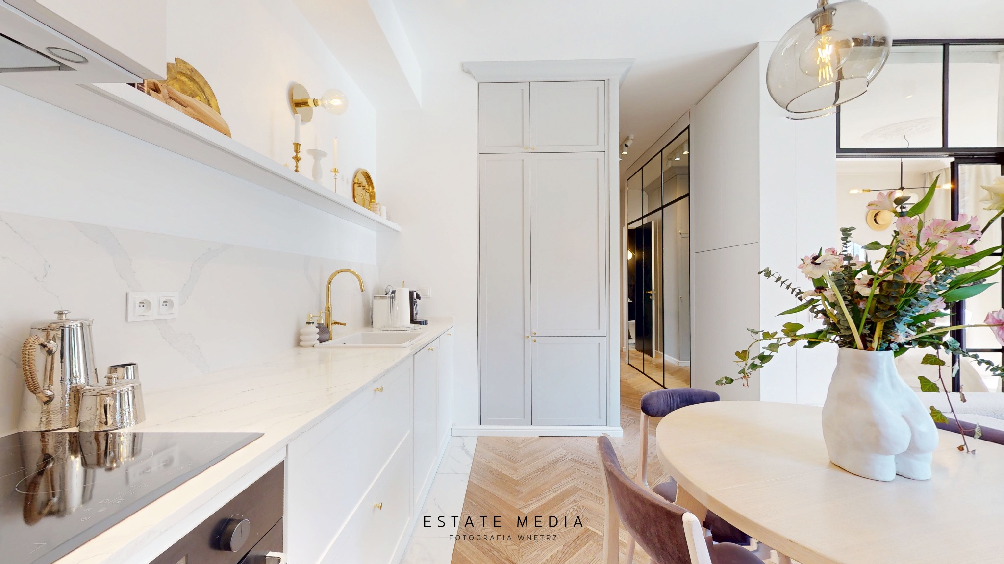 Jasna kuchnia z białymi szafkami, złotą armaturą i marmurowym blatem, uzupełniona o stół jadalny z ceramicznym wazonem w kształcie kobiecego ciała i bukietem kwiatów.