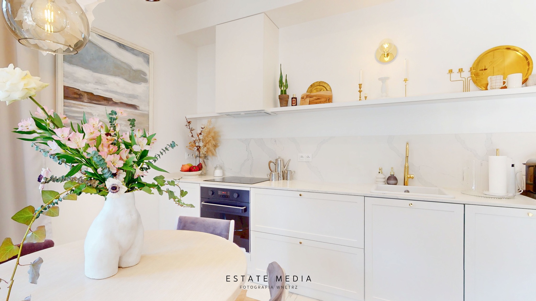 Jasna kuchnia z białymi szafkami, marmurowym blatem i dekoracjami, wazon z kwiatami na pierwszym planie.