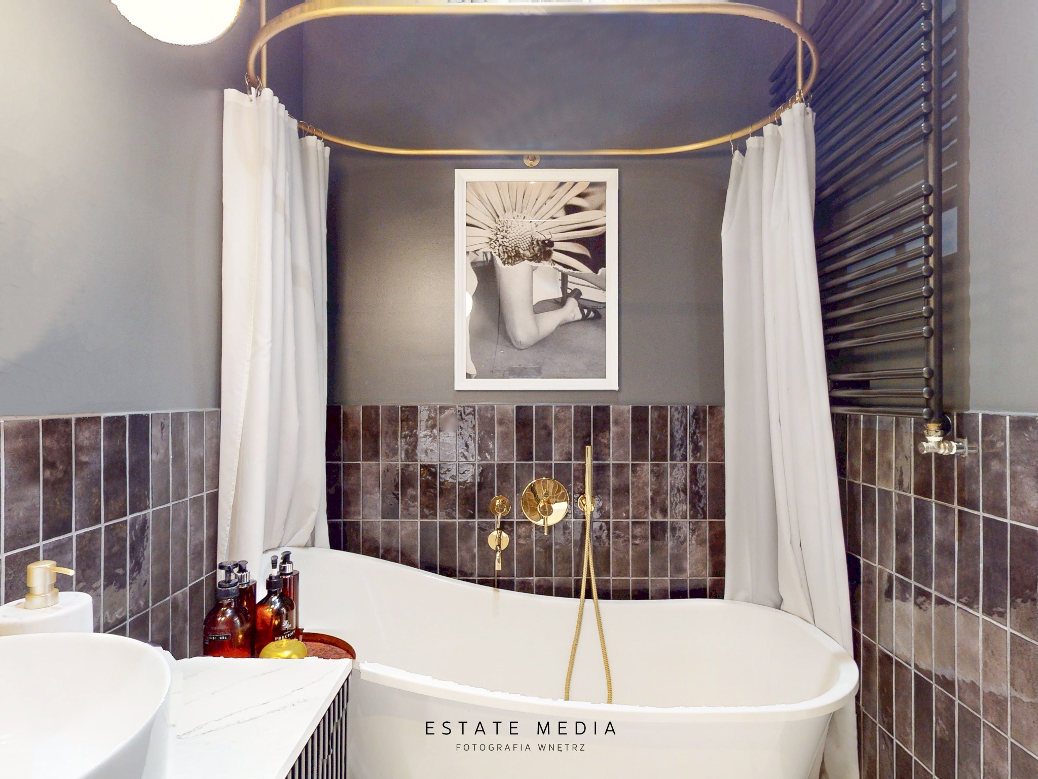 Elegancka łazienka z wolnostojącą wanną, złotą armaturą i czarno-białym zdjęciem na ścianie, oświetlona ciepłym światłem.