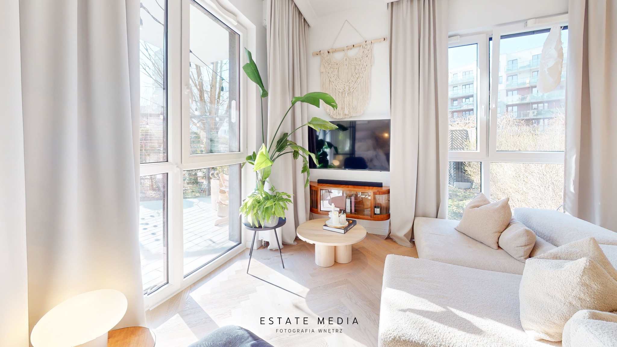 Jasne wnętrze salonu z dużym oknem, beżowymi zasłonami, sofą z poduszkami, stolikiem kawowym i roślinami doniczkowymi, oświetlone naturalnym światłem.