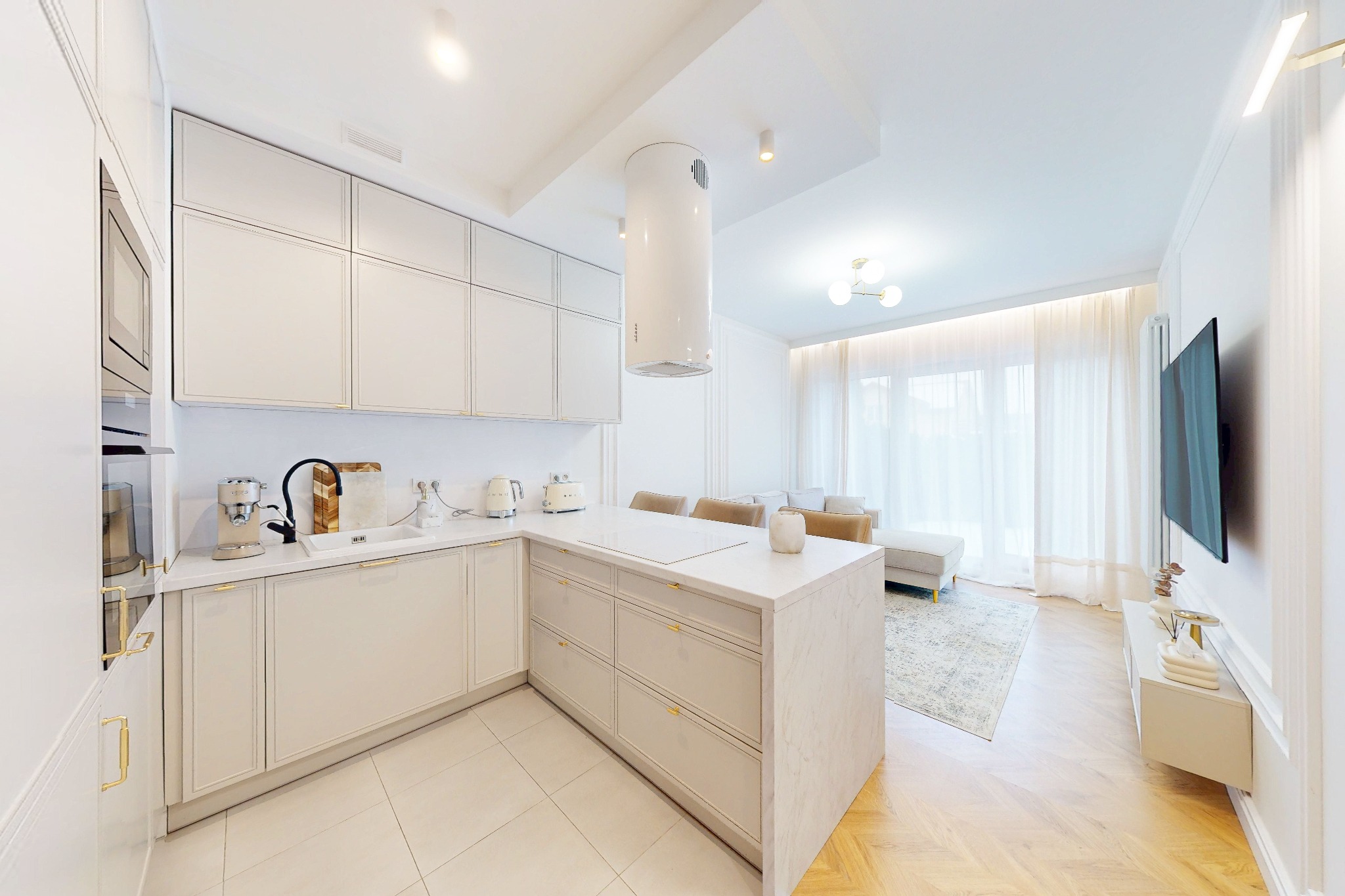 Jasna, nowoczesna kuchnia z wyspą i salonem w tle. Minimalistyczny design, białe meble z subtelnymi złotymi akcentami. Widok na salon z sofą i telewizorem.