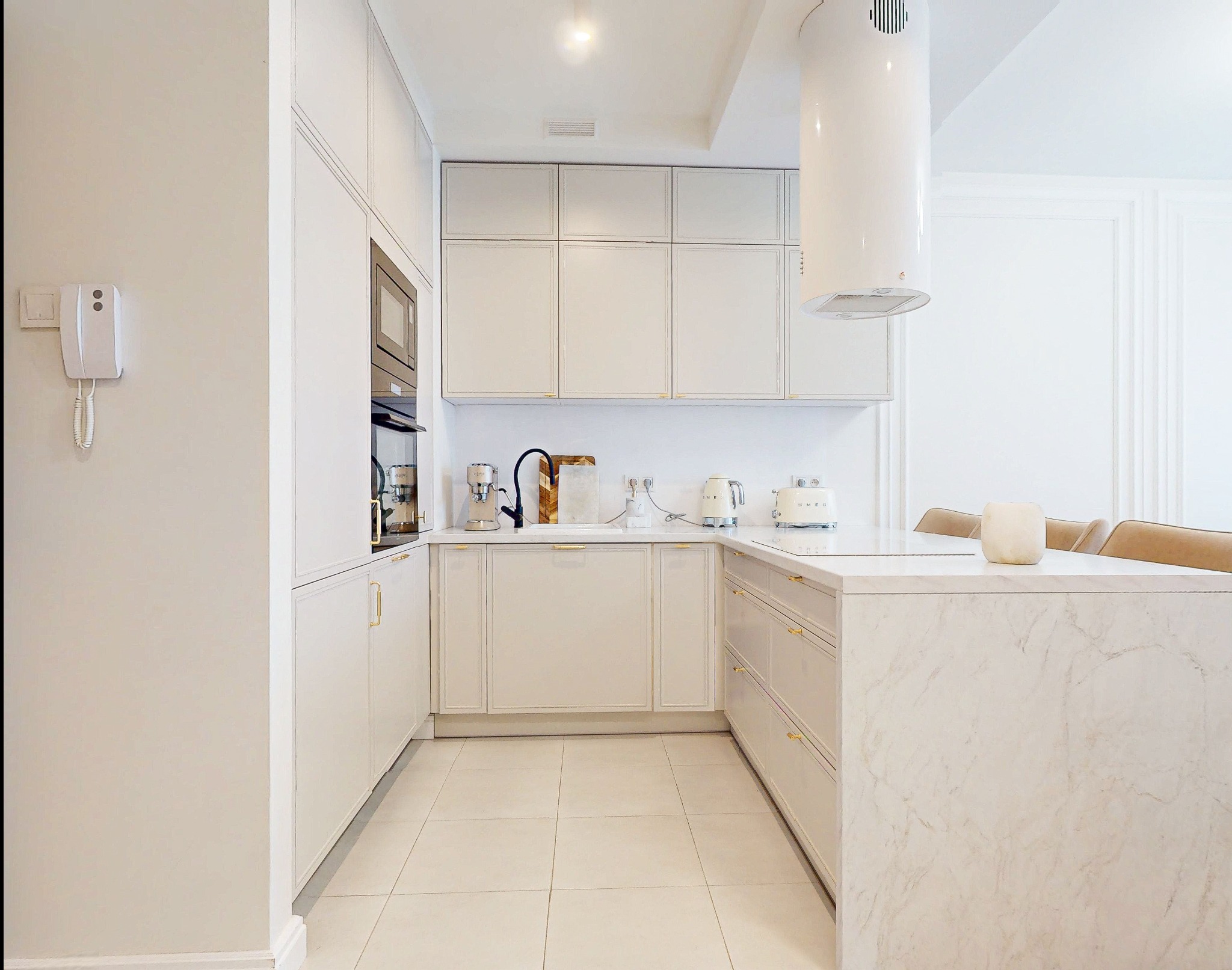 Elegancka, jasna kuchnia w stylu minimalistycznym. Białe szafki z delikatnymi złotymi uchwytami, marmurowy blat i nowoczesny okap cylindryczny. Ujęcie z perspektywy wnętrza.