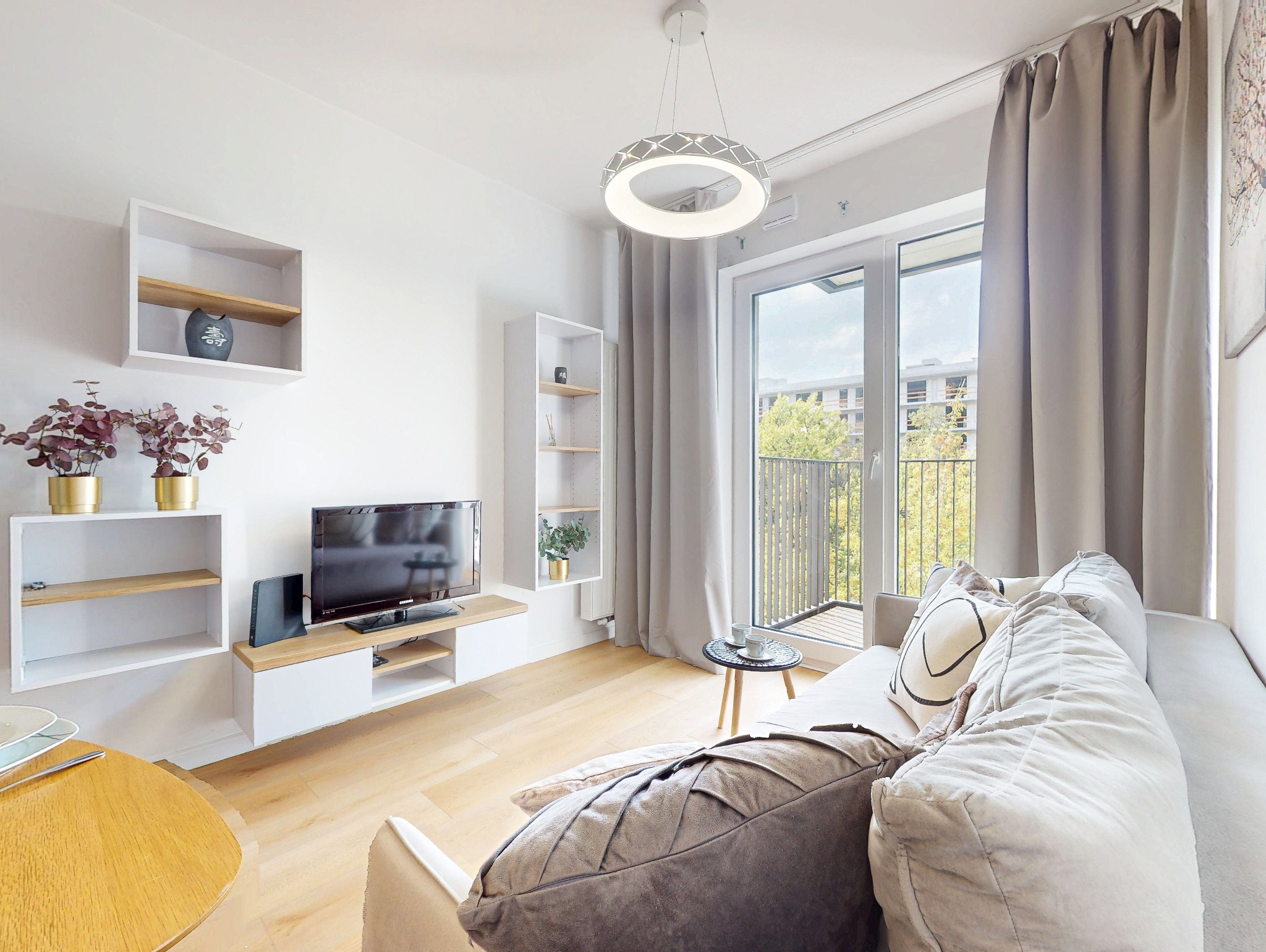 Jasne wnętrze salonu z balkonem: sofa z poduszkami, telewizor na szafce RTV, półki wiszące, okrągła lampa sufitowa. Widok na zieleń za oknem.