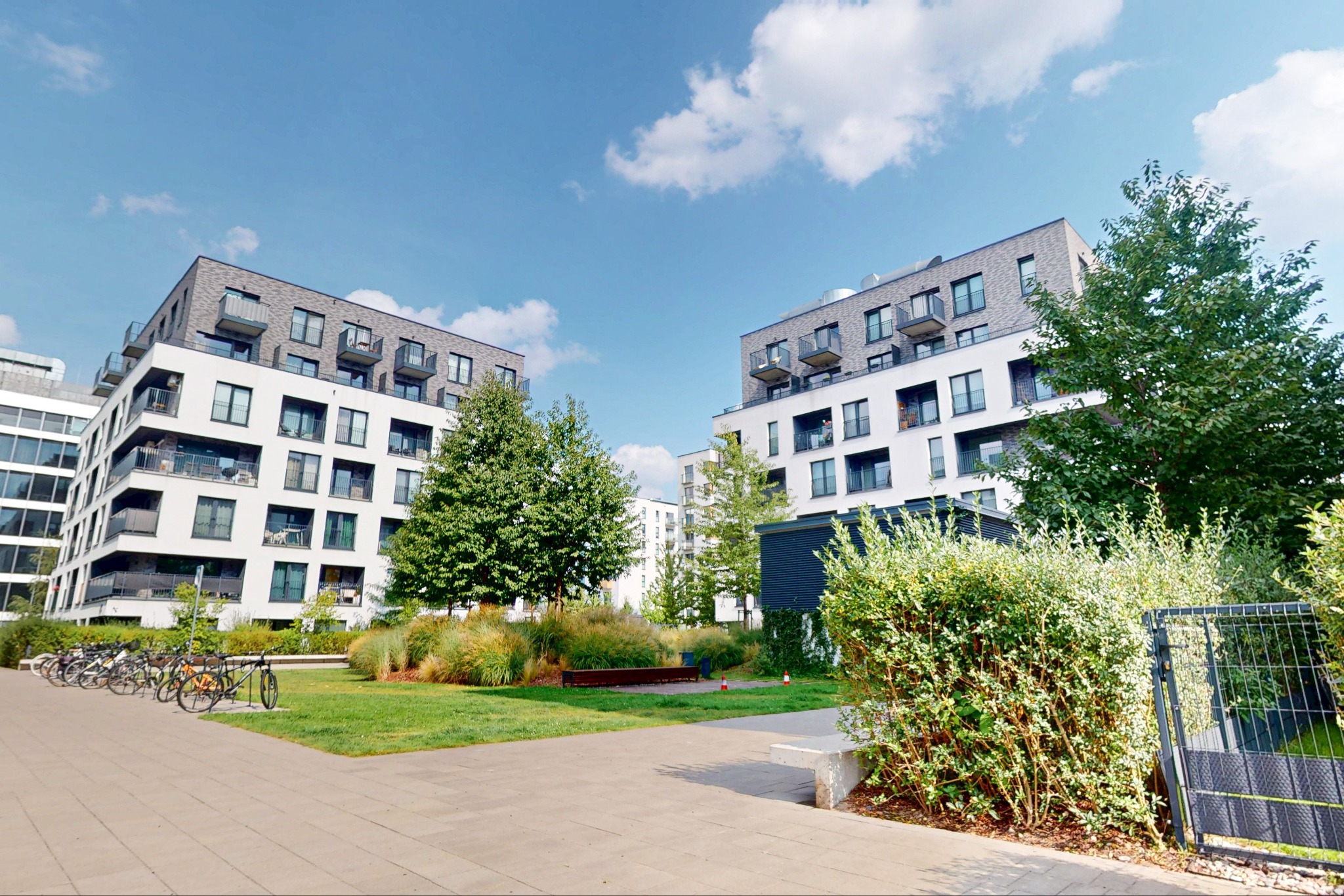 Nowoczesne budynki mieszkalne z balkonami, otoczone zielenią i ścieżką rowerową, sfotografowane w słoneczny dzień z perspektywy chodnika.