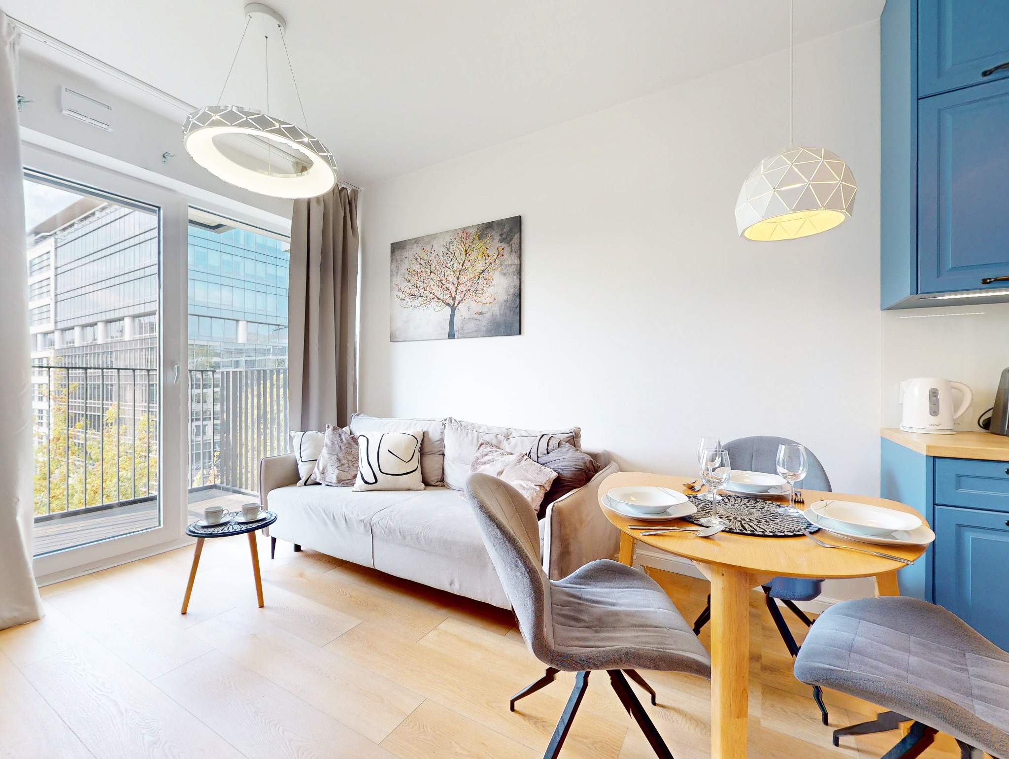 Jasne wnętrze mieszkania z drewnianą podłogą, sofą z poduszkami, okrągłym stołem z zastawą i widokiem na nowoczesny budynek zza balkonu.