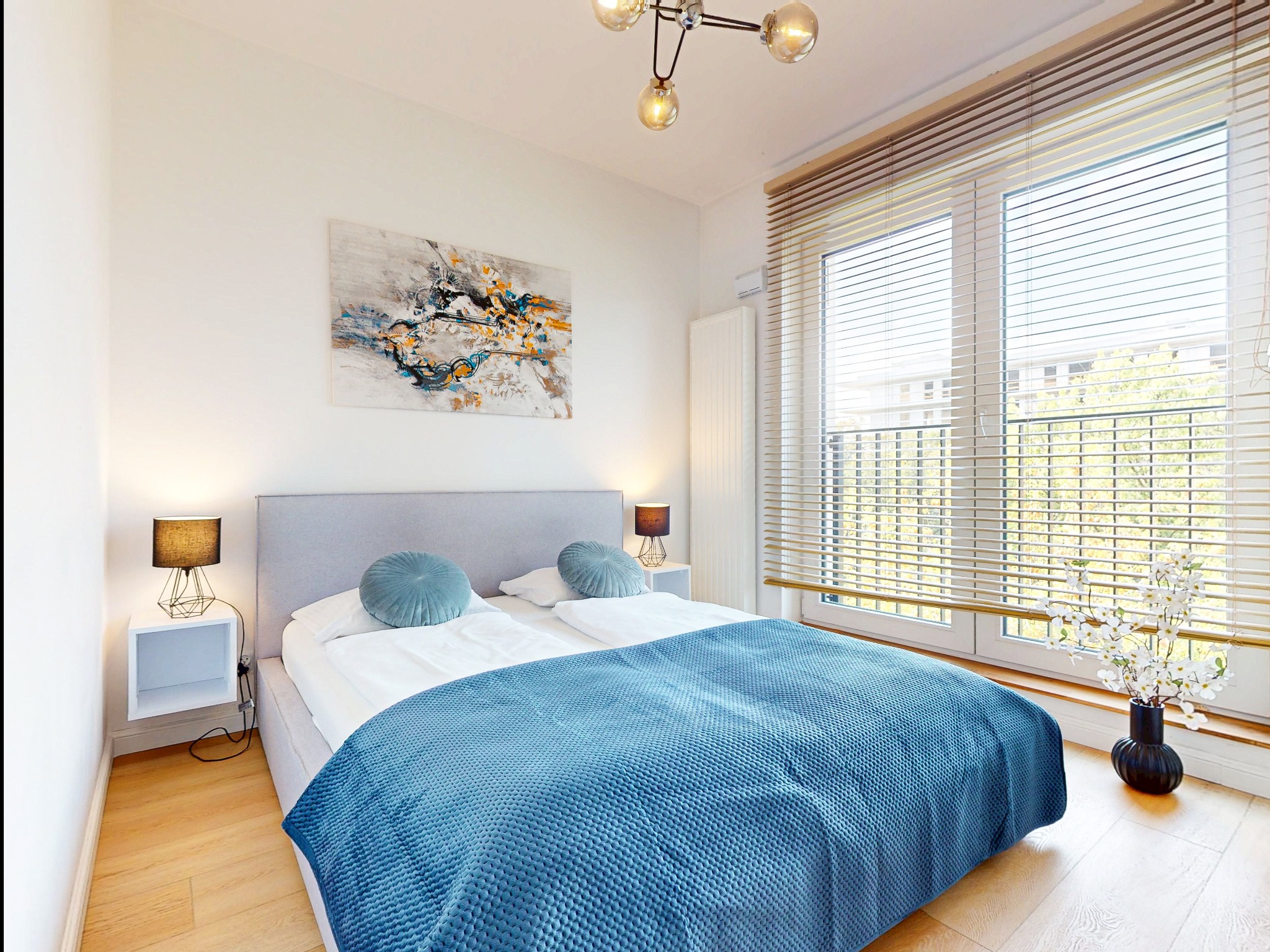 Jasna sypialnia z łóżkiem, niebieską narzutą i okrągłymi poduszkami. Na ścianie abstrakcyjny obraz, a na podłodze wazon z kwitnącymi gałązkami.