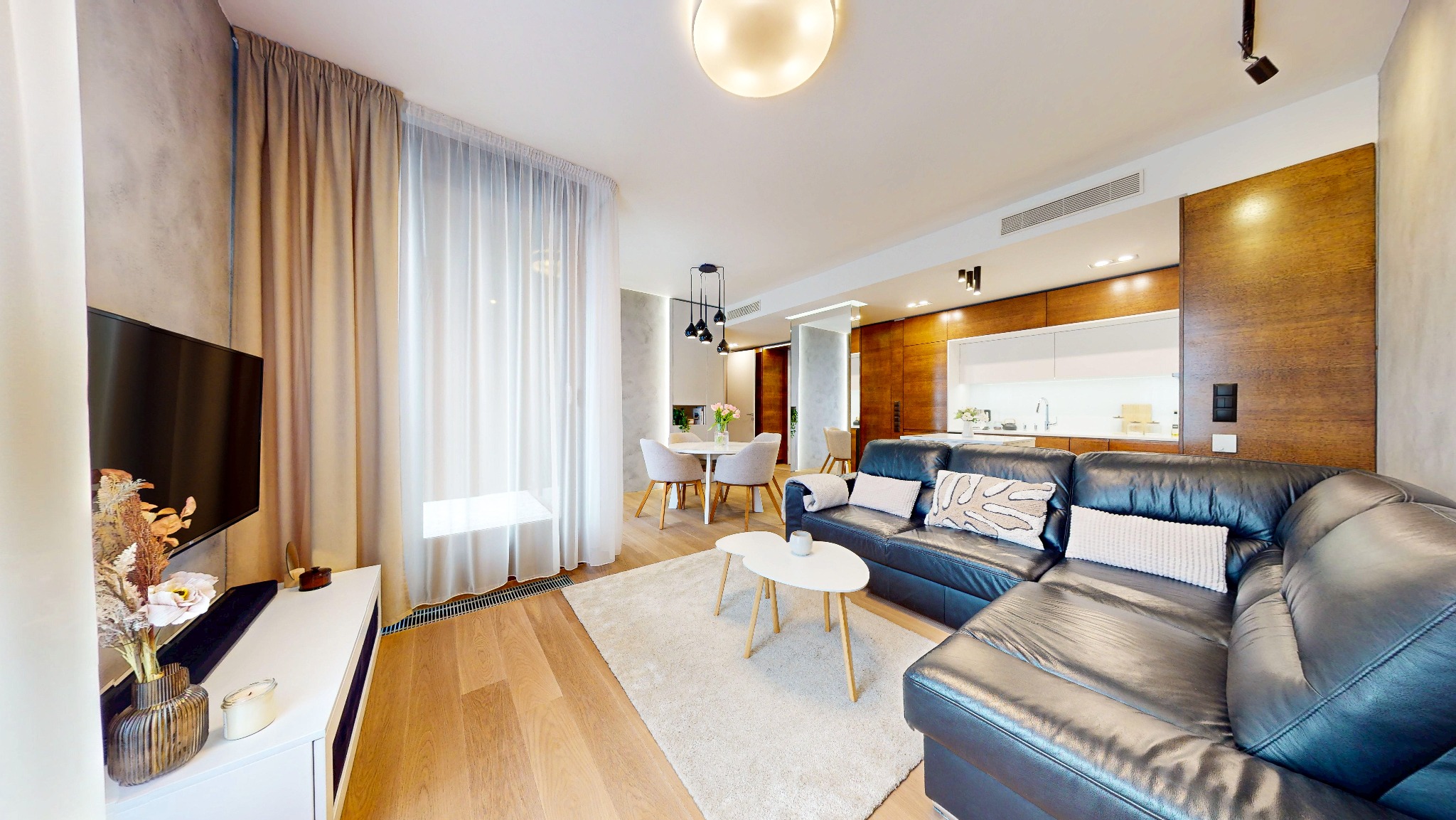 Jasne wnętrze salonu z aneksem kuchennym, skórzana sofa narożna, drewniana podłoga, okno z zasłonami, stół z krzesłami. Nowoczesny design, ciepłe oświetlenie.