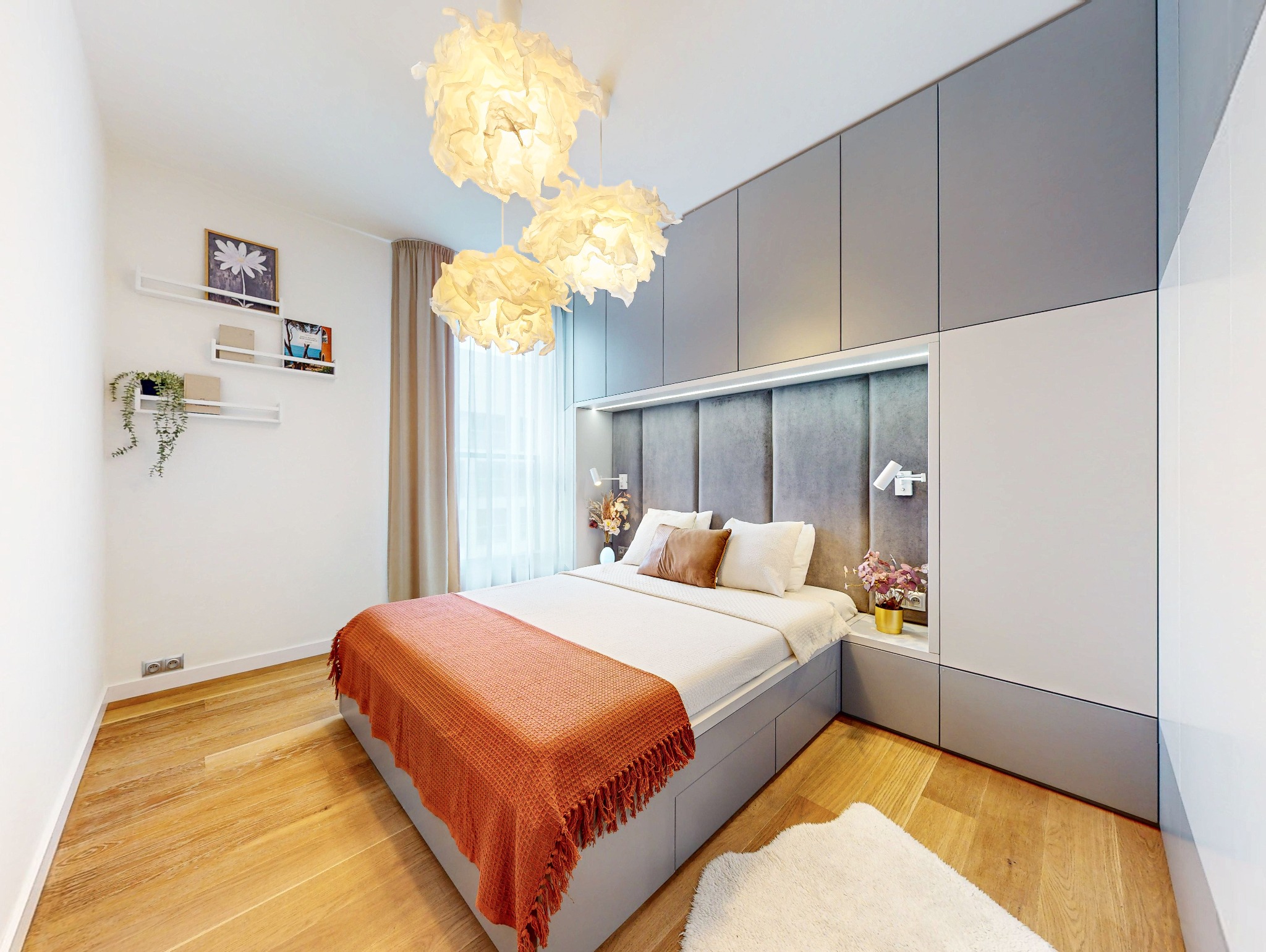 Nowoczesna sypialnia z drewnianą podłogą, szarą zabudową i dekoracyjnym oświetleniem. Pomarańczowy pled na łóżku dodaje ciepła wnętrzu.
