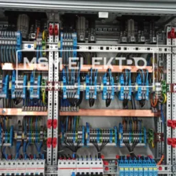 Rozbudowana skrzynka elektryczna z licznymi niebieskimi złączkami, miedzianymi szynami i przewodami w różnych kolorach, zamontowana w metalowej obudowie z widocznymi elementami mocującymi.