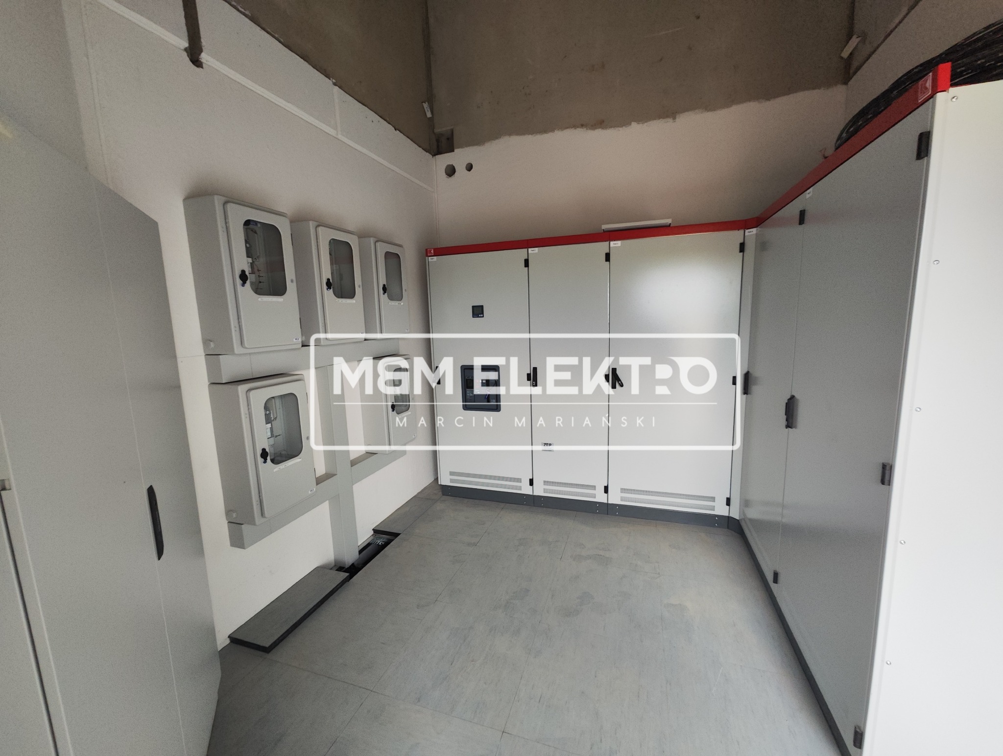 Wnętrze pomieszczenia z zainstalowanymi rozdzielnicami elektrycznymi i licznikami, widoczne kable w górnej części kadru, szara podłoga i surowy betonowy sufit.