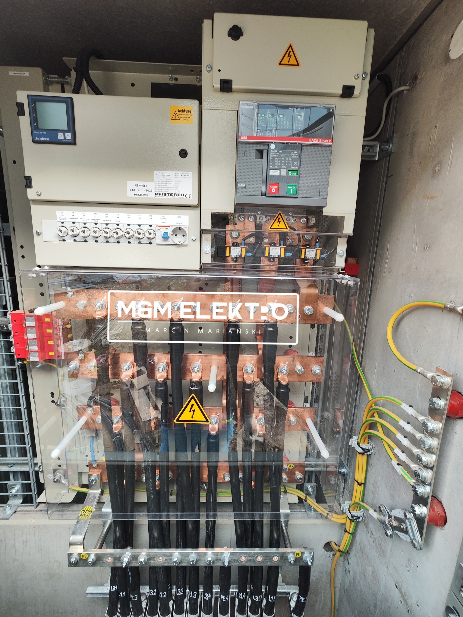 Rozdzielnica elektryczna z widocznymi bezpiecznikami, przewodami oznaczonymi numerami, logiem firmy MGM ELEKTRO i tabliczkami ostrzegawczymi o napięciu, zamontowana w szarej obudowie.