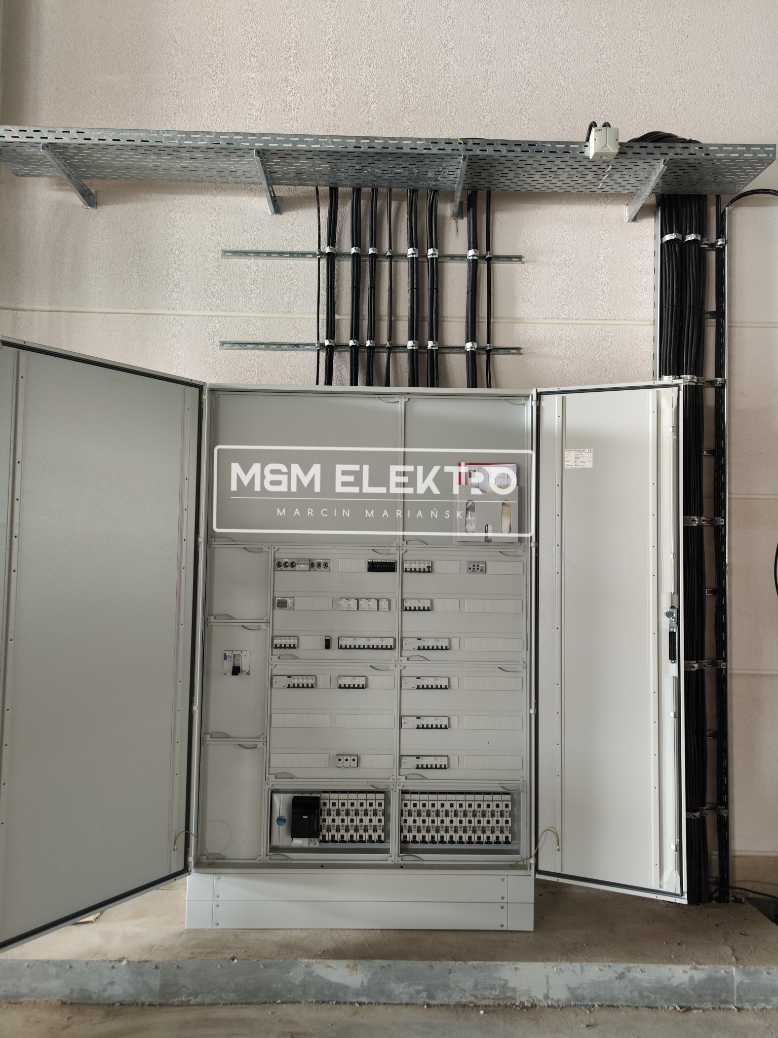 Otwarta szafa elektryczna z widocznymi zabezpieczeniami i okablowaniem, kable prowadzone w korytach na ścianie, widoczne logo firmy M&M Elektro Marcin Mariański.