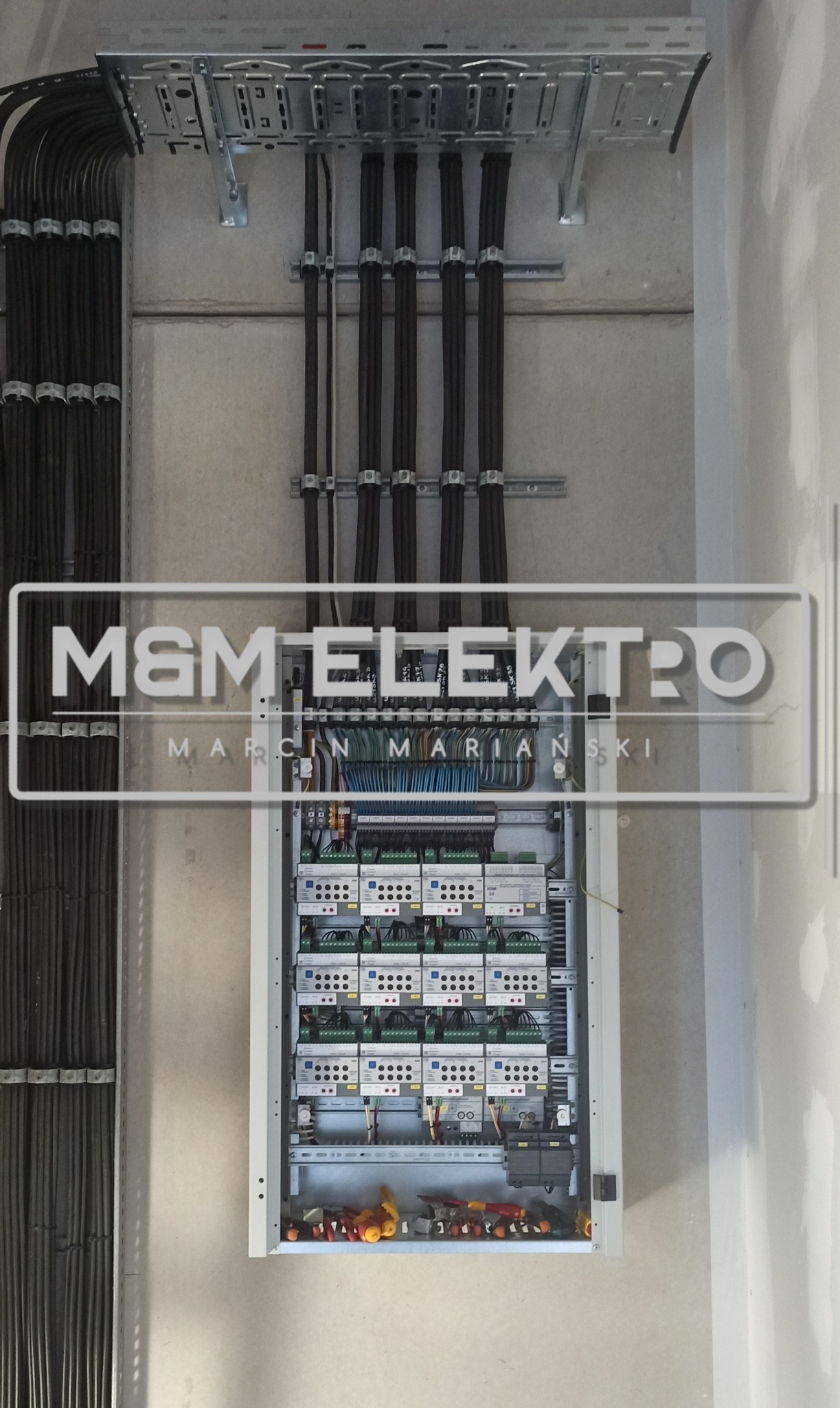 Rozdzielnia elektryczna z licznymi przewodami ułożonymi w korytkach, widoczne zabezpieczenia i moduły sterujące, logo firmy na pierwszym planie.