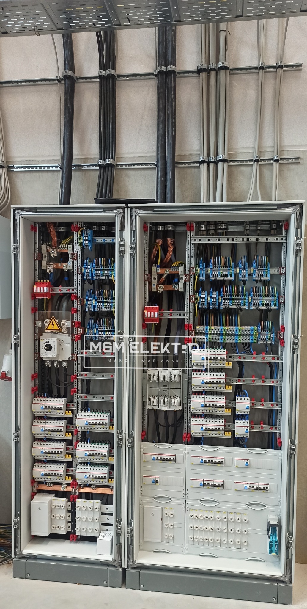 Rozdzielnica elektryczna dwudrzwiowa z widocznymi bezpiecznikami, przewodami i elementami sterującymi, zamontowana na ścianie z widocznymi kablami w korytach