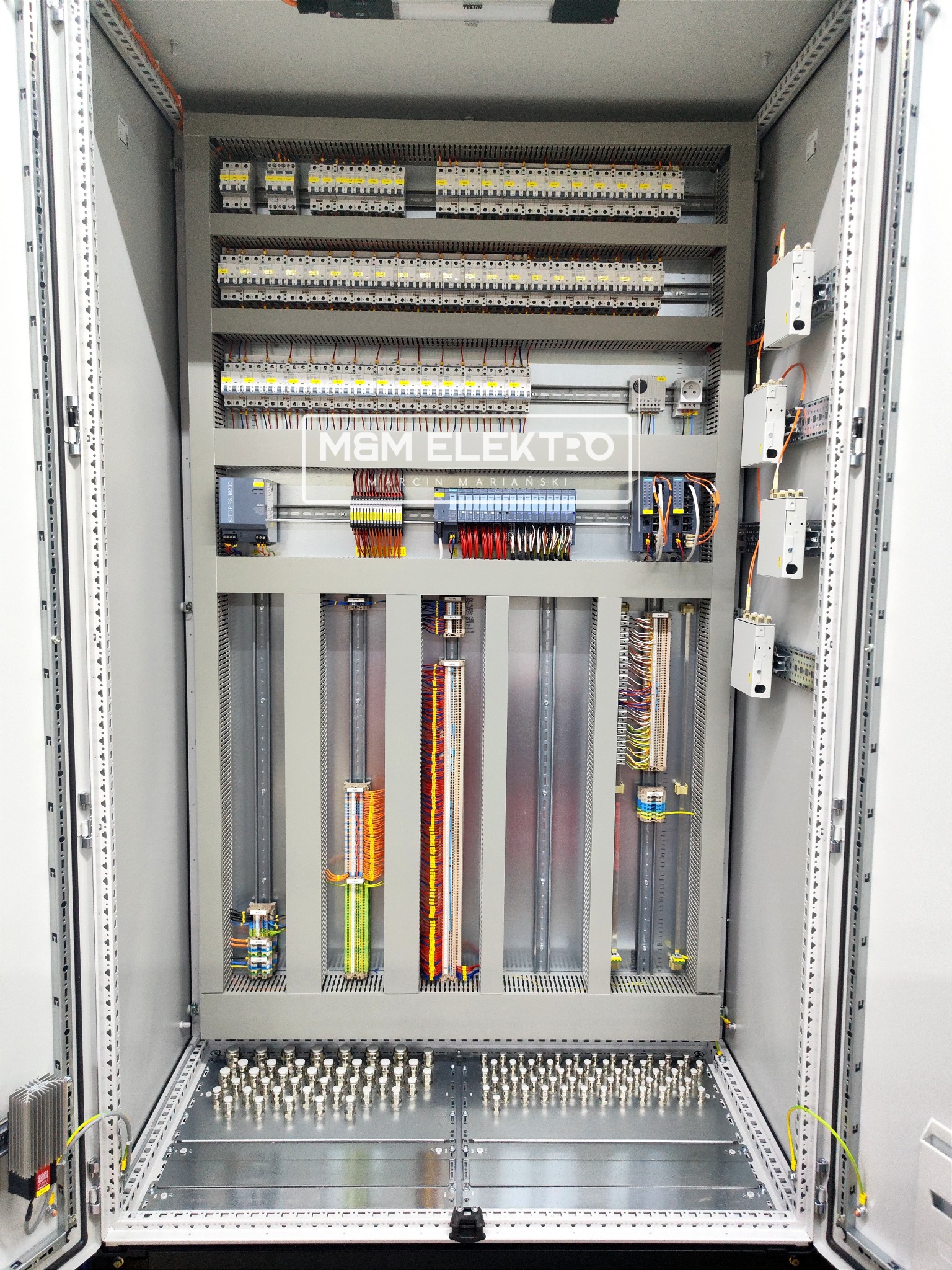 Otwarta szafa sterownicza z widocznymi zabezpieczeniami, modułami PLC, okablowaniem i listwami zaciskowymi, zainstalowana w pomieszczeniu.