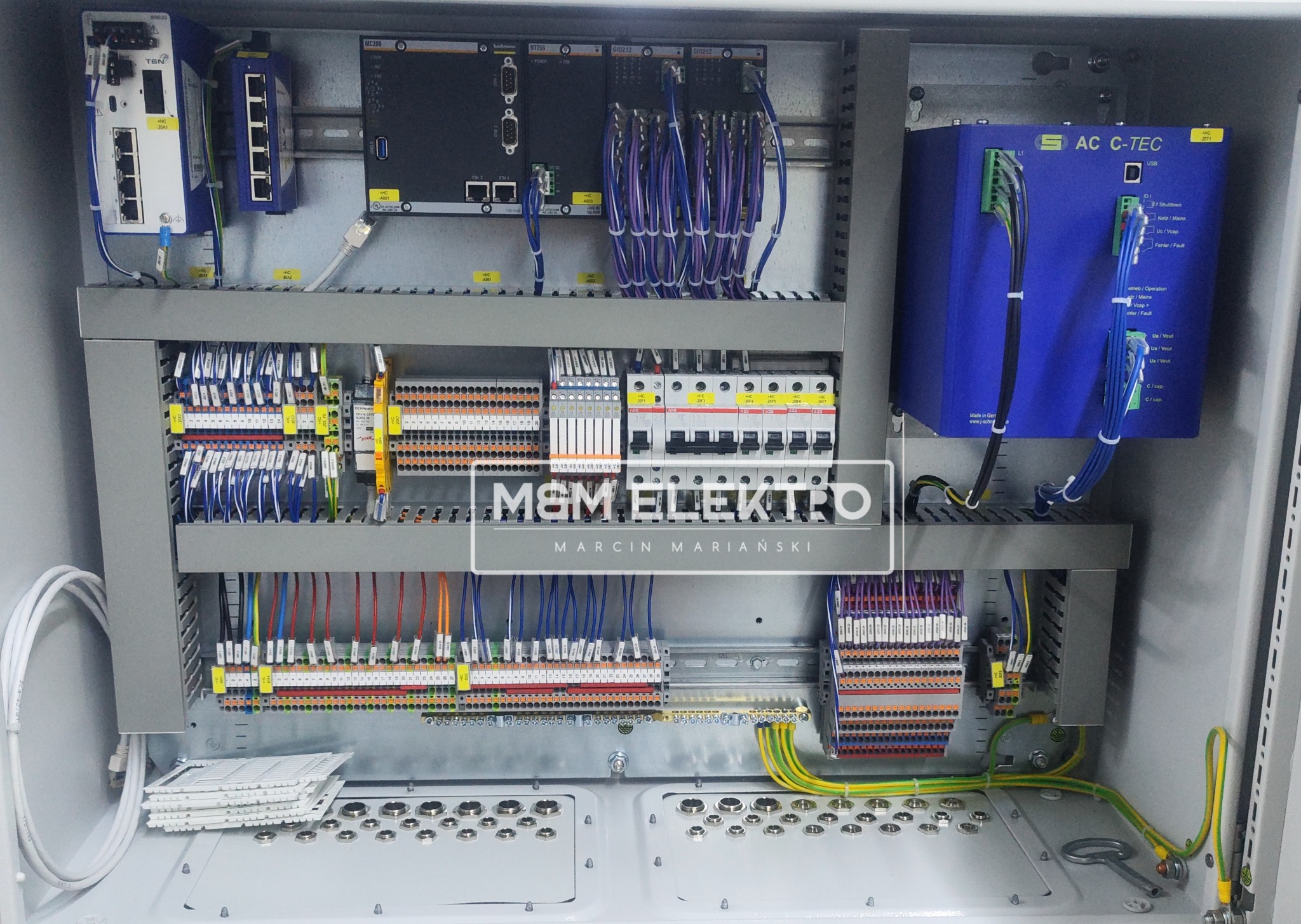 Otwarta szafa sterownicza z widocznymi modułami, kablami i złączkami, uporządkowana instalacja elektryczna w szarej obudowie, z elementami sieciowymi i zasilającymi, w tle logo firmy M&M Elektro.