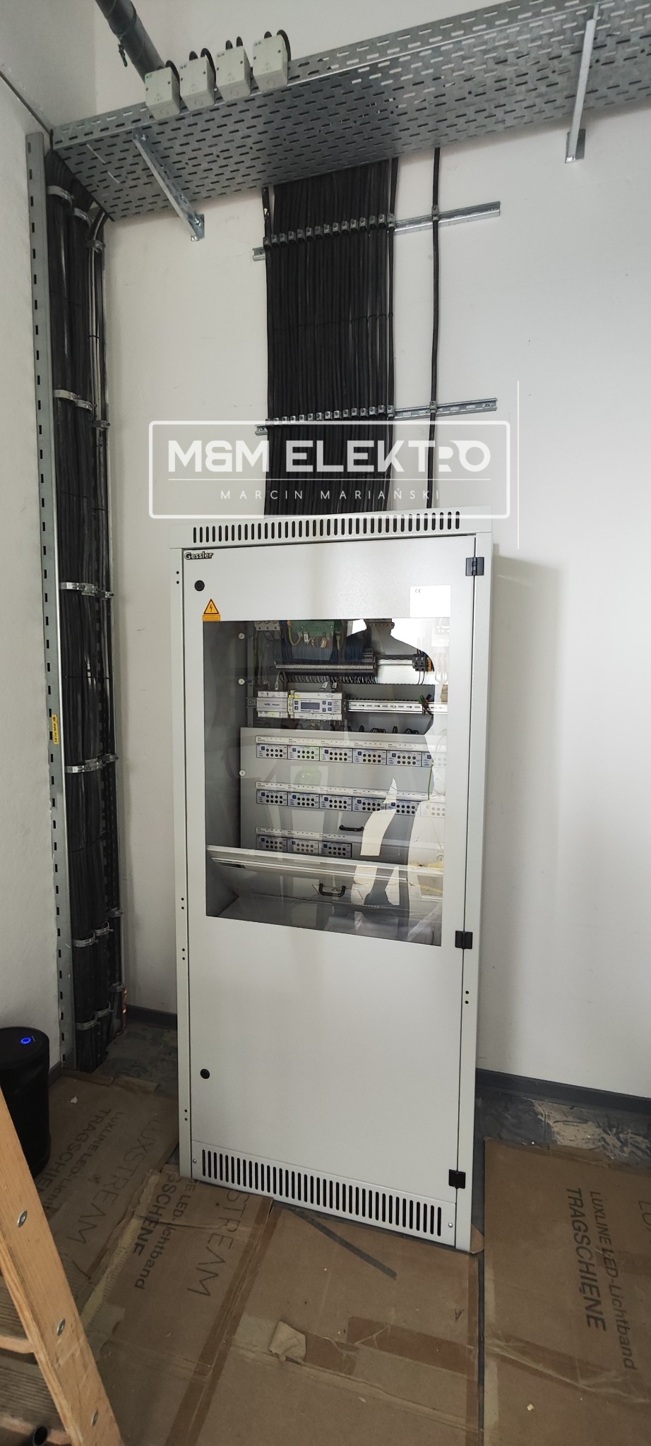 Szafa sterownicza z widocznymi elementami elektrycznymi wewnątrz, kable prowadzone w korytach i po ścianie, logo firmy nałożone na zdjęcie.
