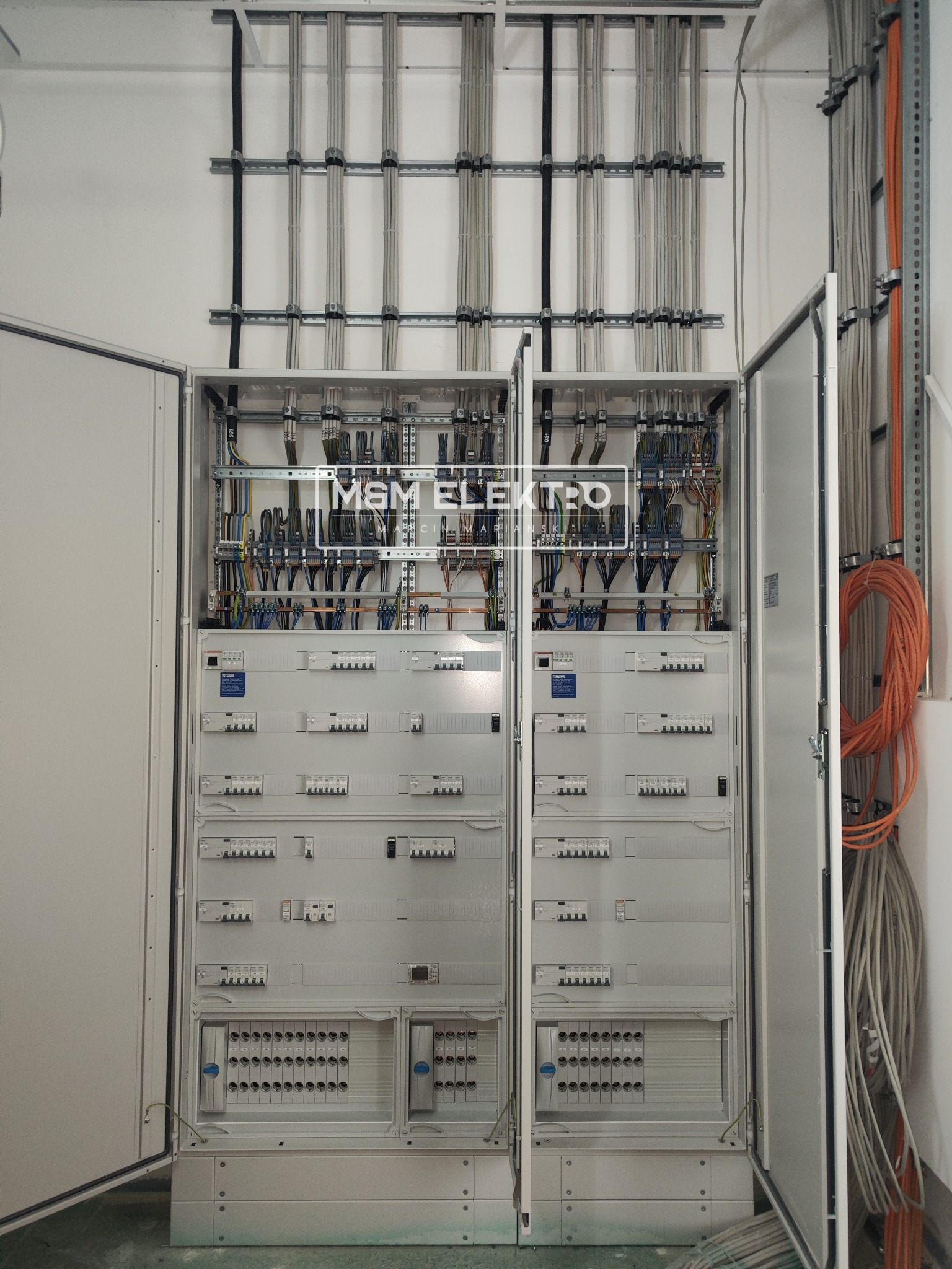 Otwarta szafa elektryczna z widocznymi bezpiecznikami, przewodami i listwami montażowymi, z logo firmy MSM ELEKTO na środku, dodatkowe kable poprowadzone w korytach na ścianie.