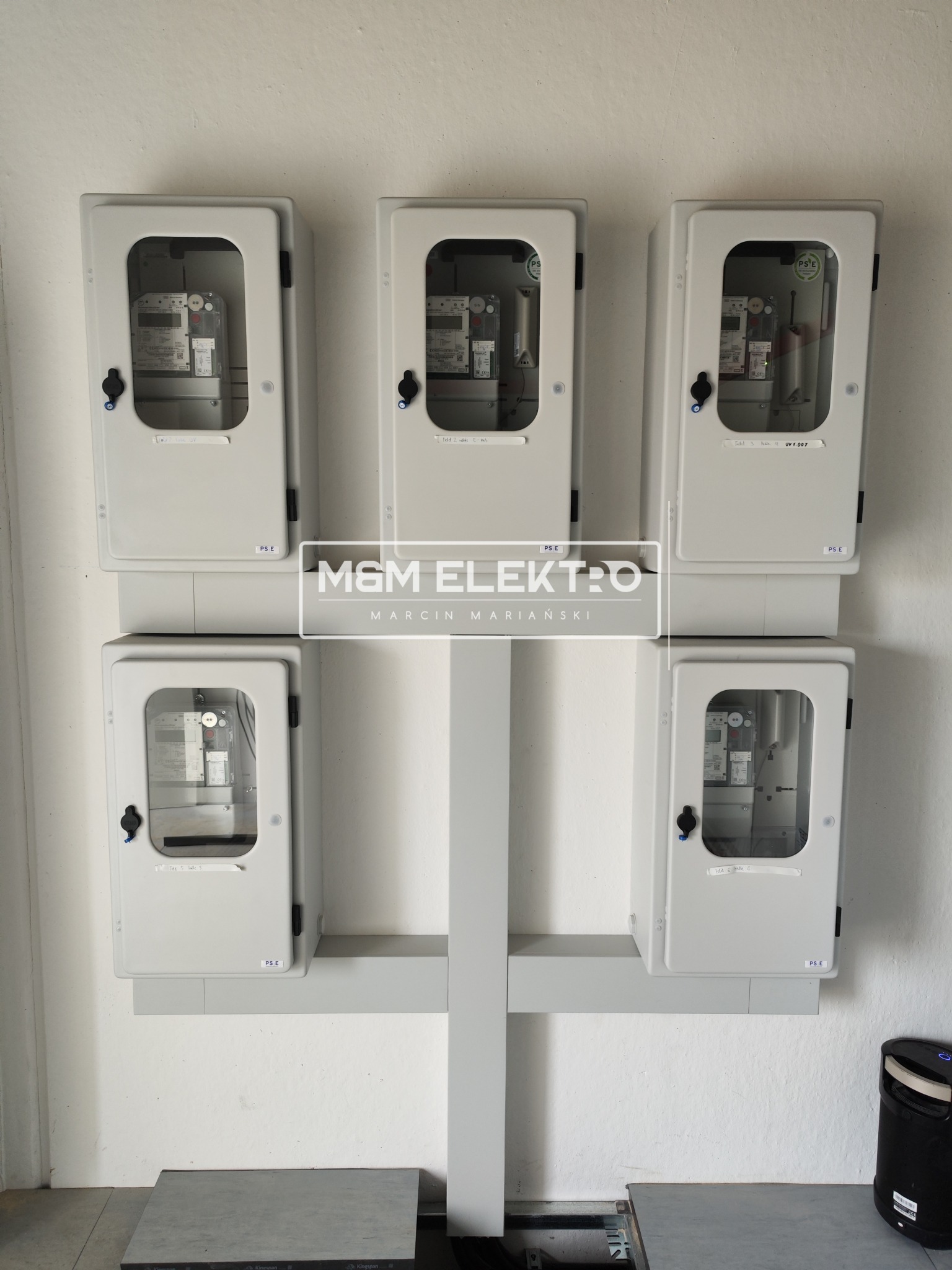 Zestaw pięciu szarych, plastikowych skrzynek z przezroczystymi okienkami, zawierających liczniki elektryczne, zamontowanych na białej ścianie na szarej konstrukcji wsporczej. Widoczne etykiety...