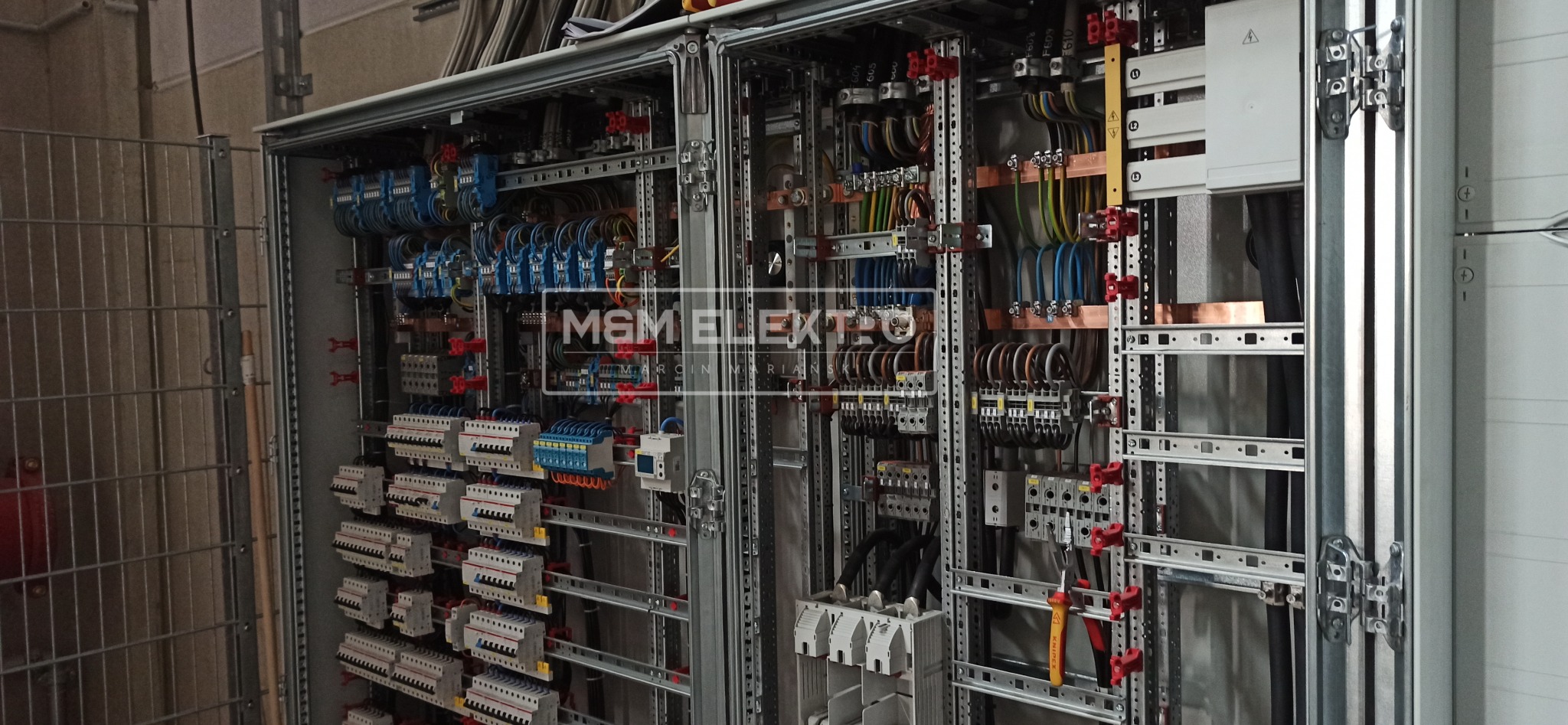 Otwarta szafa rozdzielcza z licznymi zabezpieczeniami, przewodami i listwami zaciskowymi, widoczne narzędzia elektryczne, szyny montażowe z czerwonymi uchwytami, przewody ułożone w wiązki.